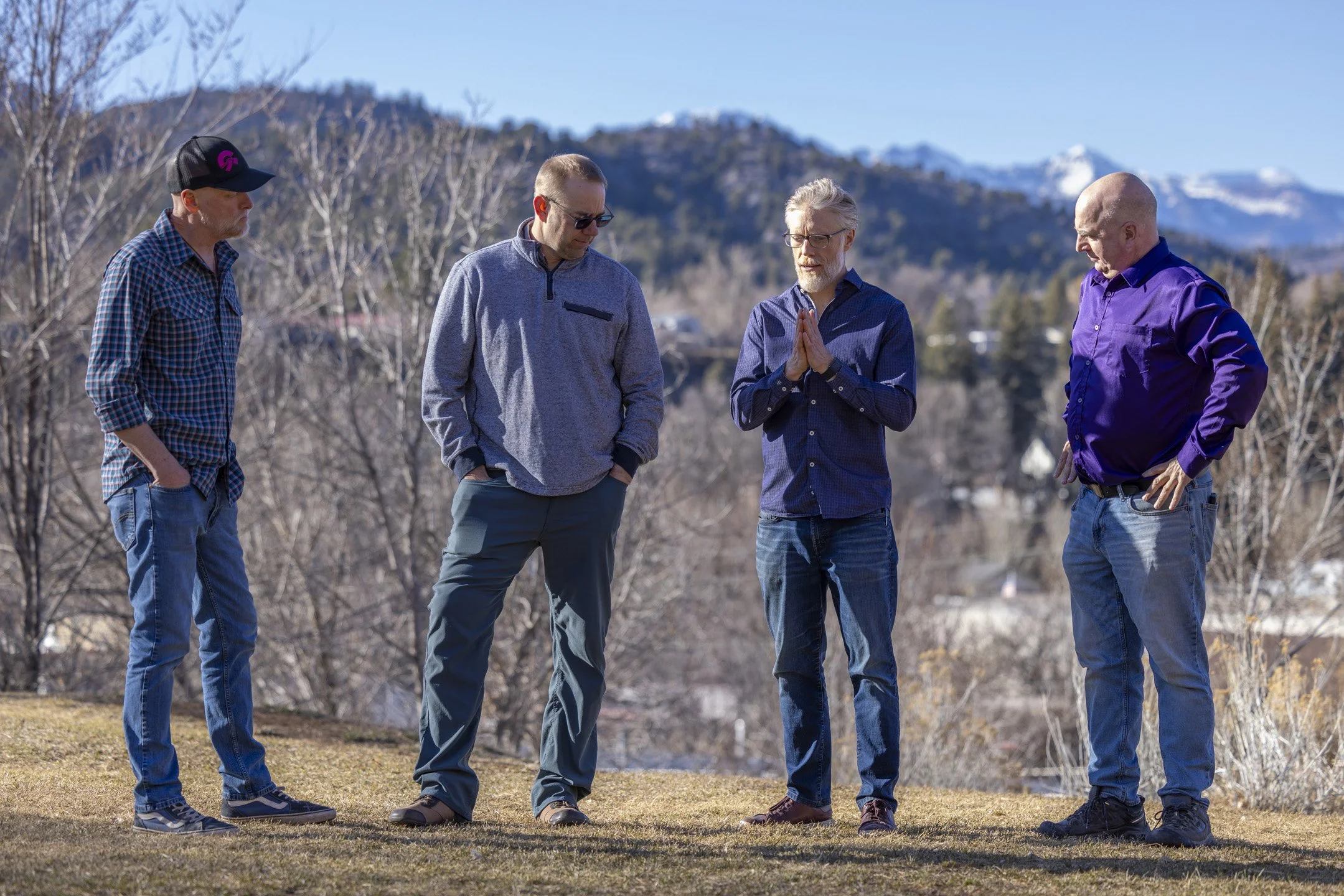 Four men standing outdoors on grass, with mountains in the background. One man is wearing a checked shirt and cap, two have hands in pockets, and one is clasping his hands.