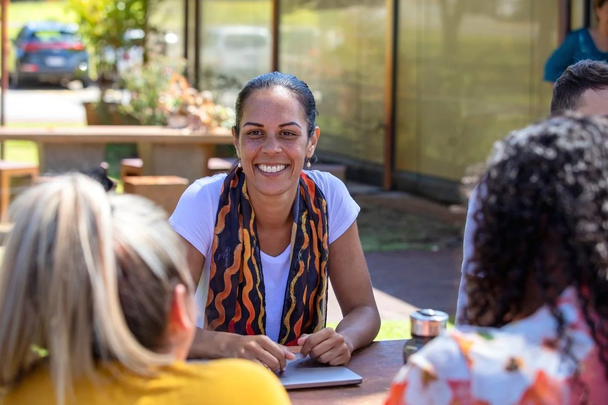 A person smiling and sitting at an outdoor table with a laptop, wearing a white shirt and a colorful scarf, surrounded by others in a casual setting.