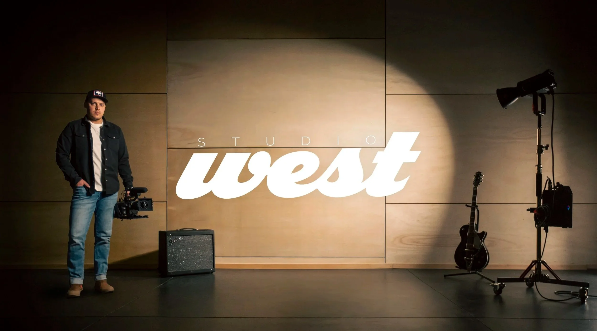 Man standing in a music studio holding a camera with the logo 'Studio West' projected on a wood-paneled wall; music equipment and lighting on stands in the background.
