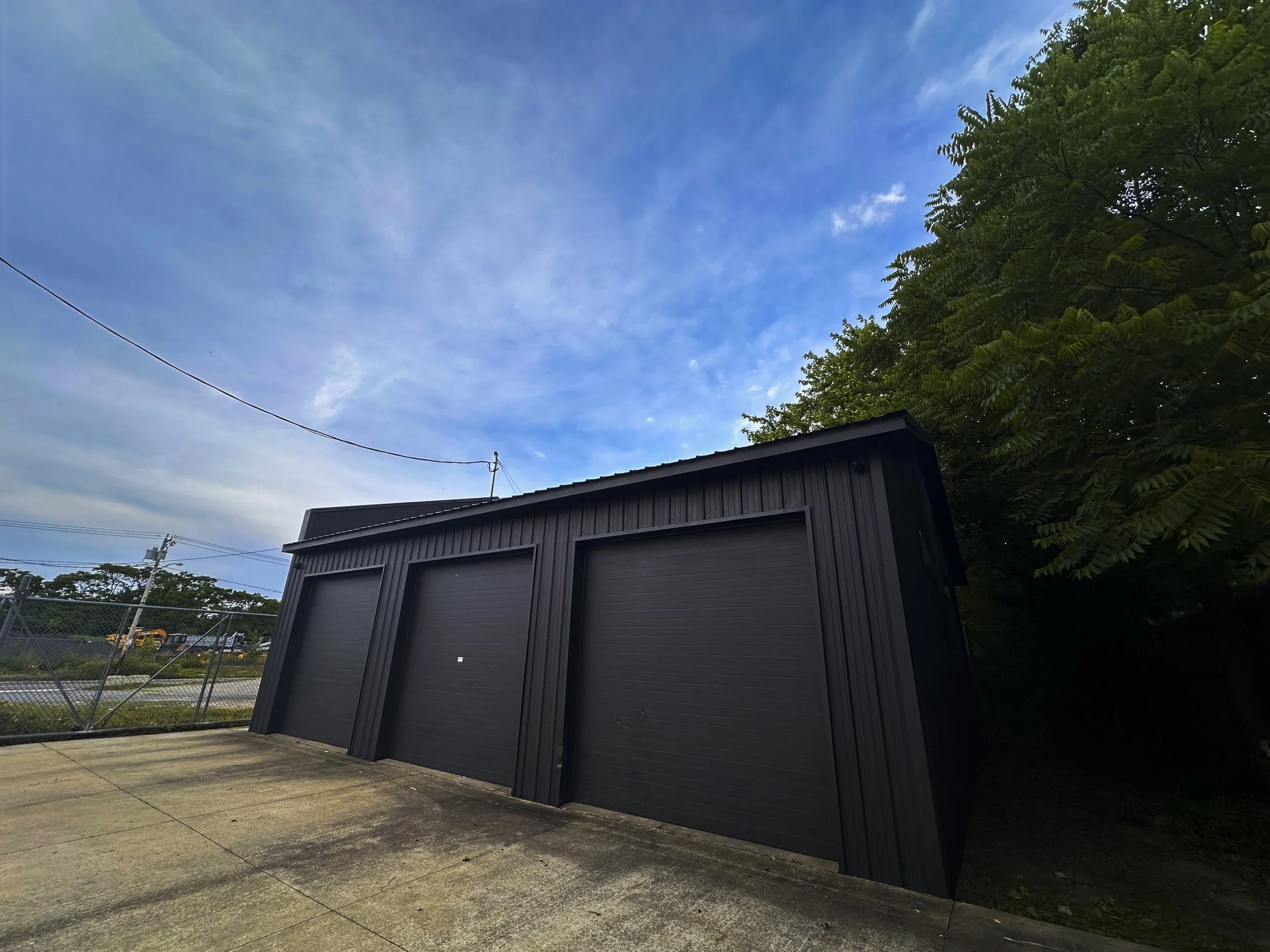 Black metal garage with three closed doors situated on a concrete driveway, with trees on the right and a fence and utility poles on the left, under a partly cloudy blue sky.