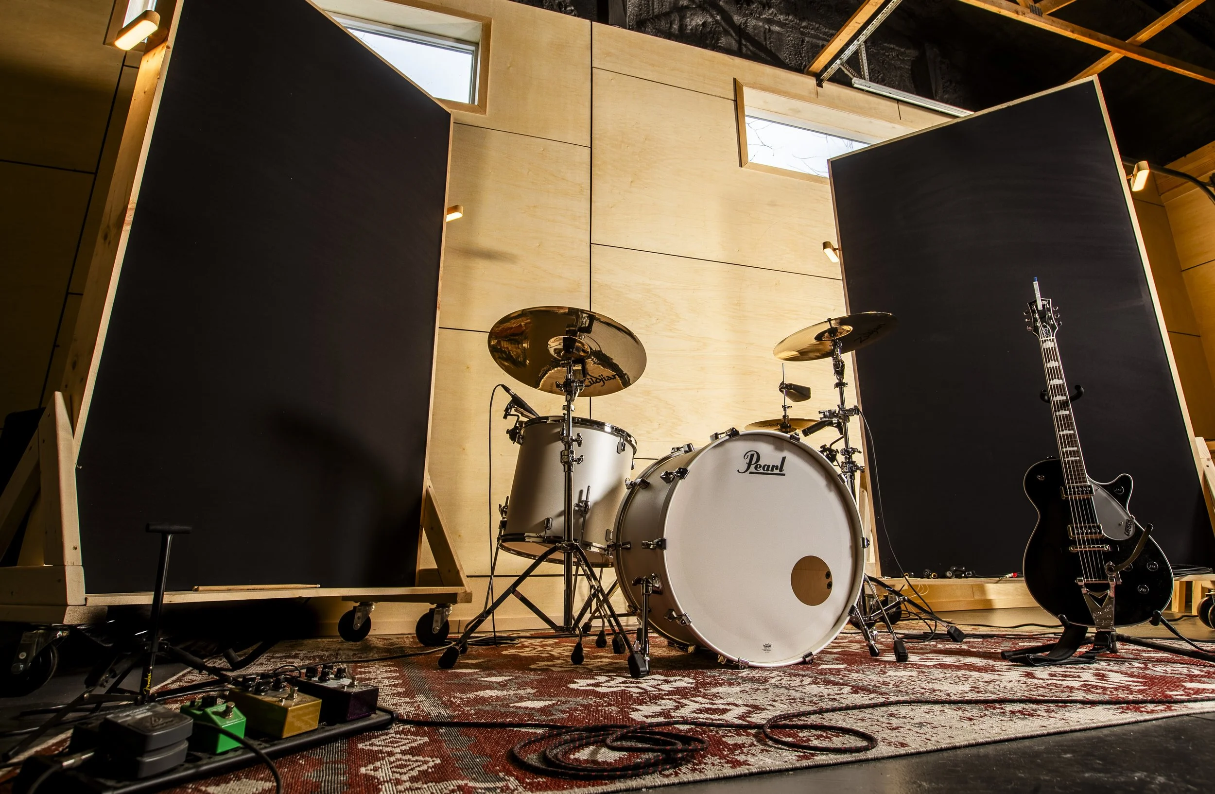 Empty drum set with two large black panels, a black electric guitar, and guitar pedals on a patterned rug in a music studio.