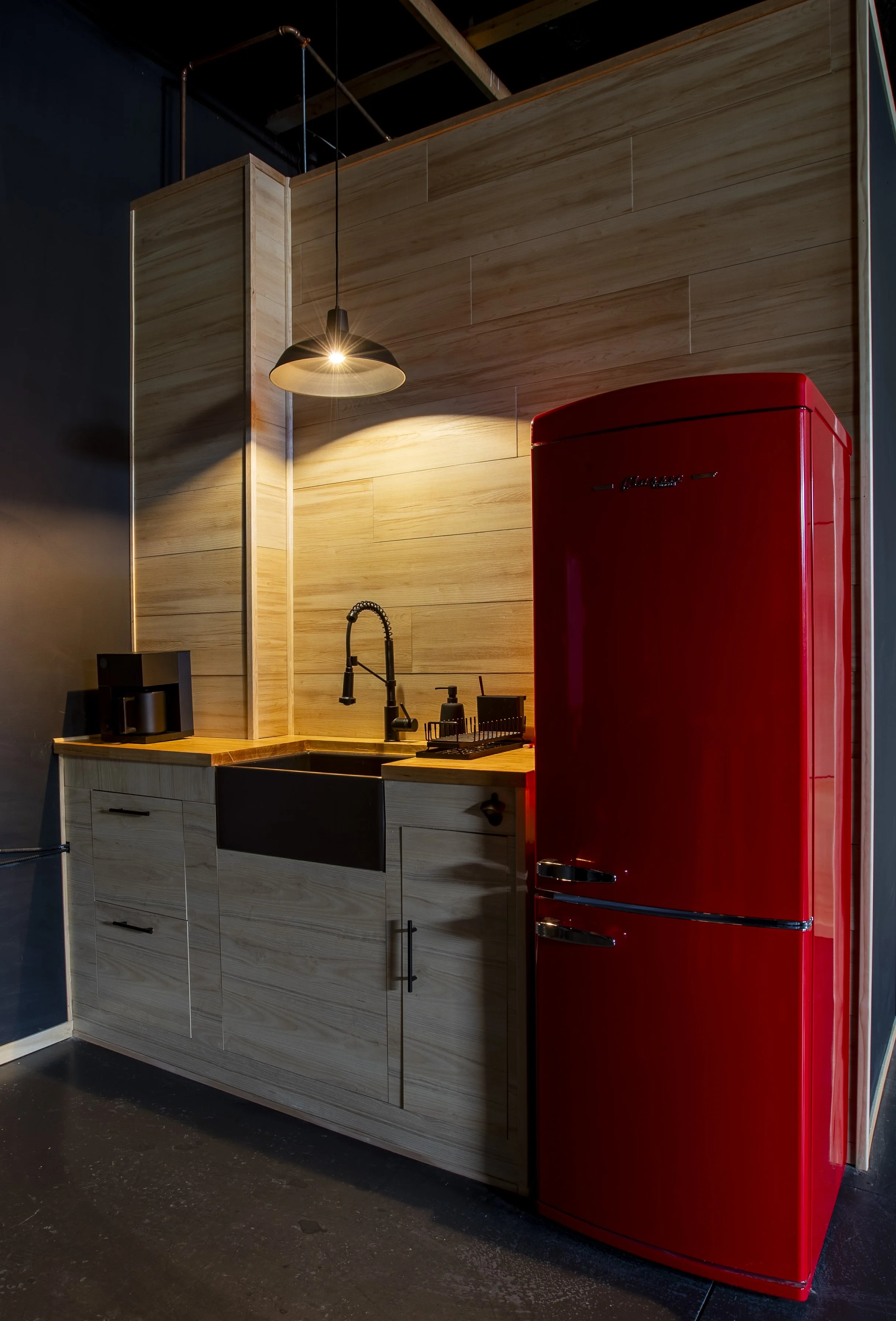 A modern kitchen corner with a wooden cabinet, black sink, black faucet, red retro-style refrigerator, and black small appliances with a black wall and an overhead piping and light fixture.