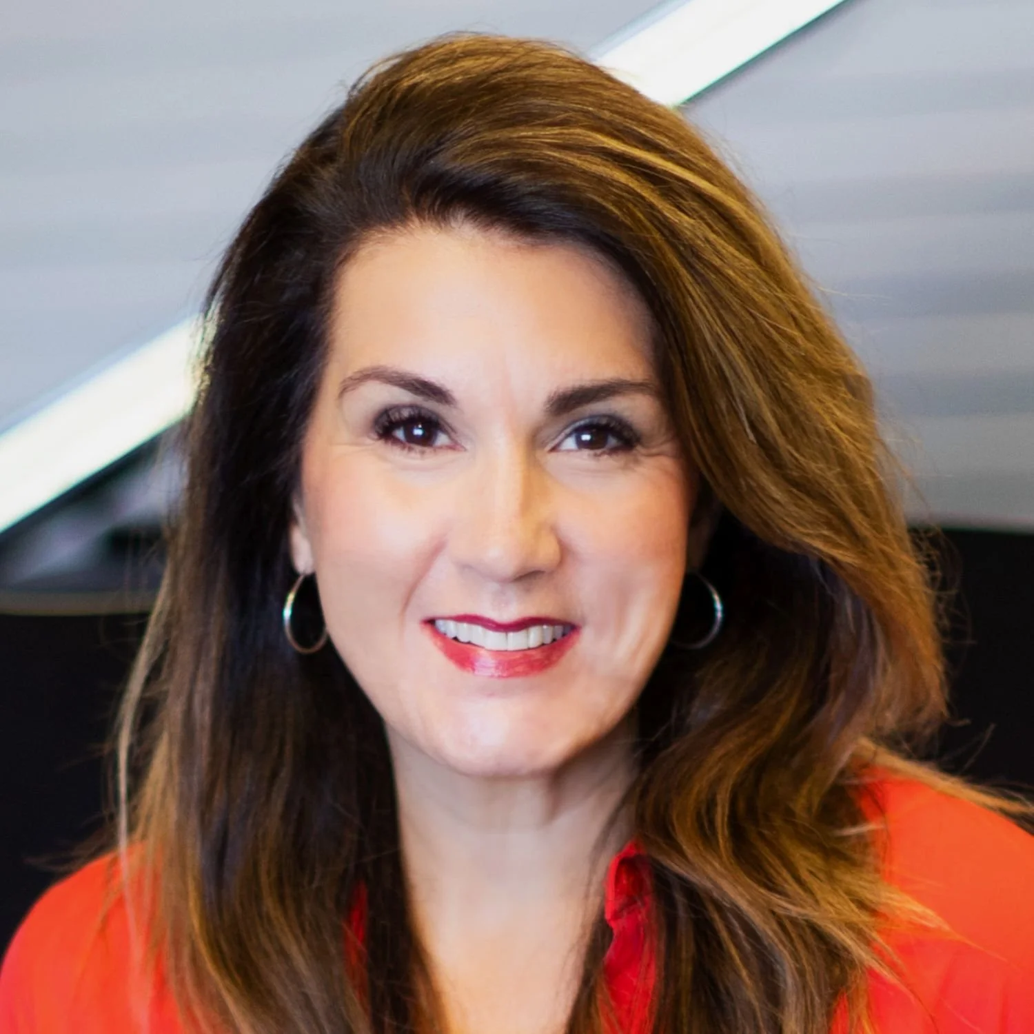 Kim Koch (3rd generation Saied Music Company) with long brown hair smiling, wearing a red blazer, earrings, and makeup, indoors with a ceiling and lights visible in the background.