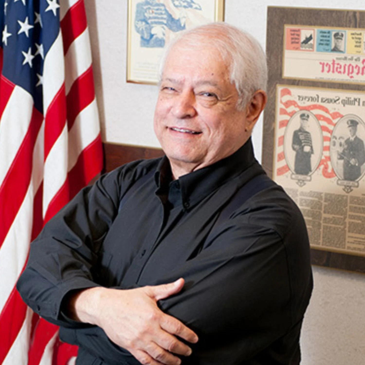 Portrait of Bob Saied (2nd generation Saied Music Company) wearing a black shirt, standing with arms crossed in front of American flags and patriotic posters on a wall.