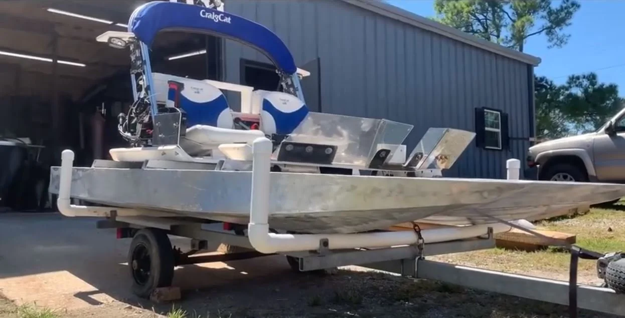 Unfinished aluminum boat on a trailer with two outboard motors and a blue canopy, parked outdoors near a building with a gray wall and a parked car.
