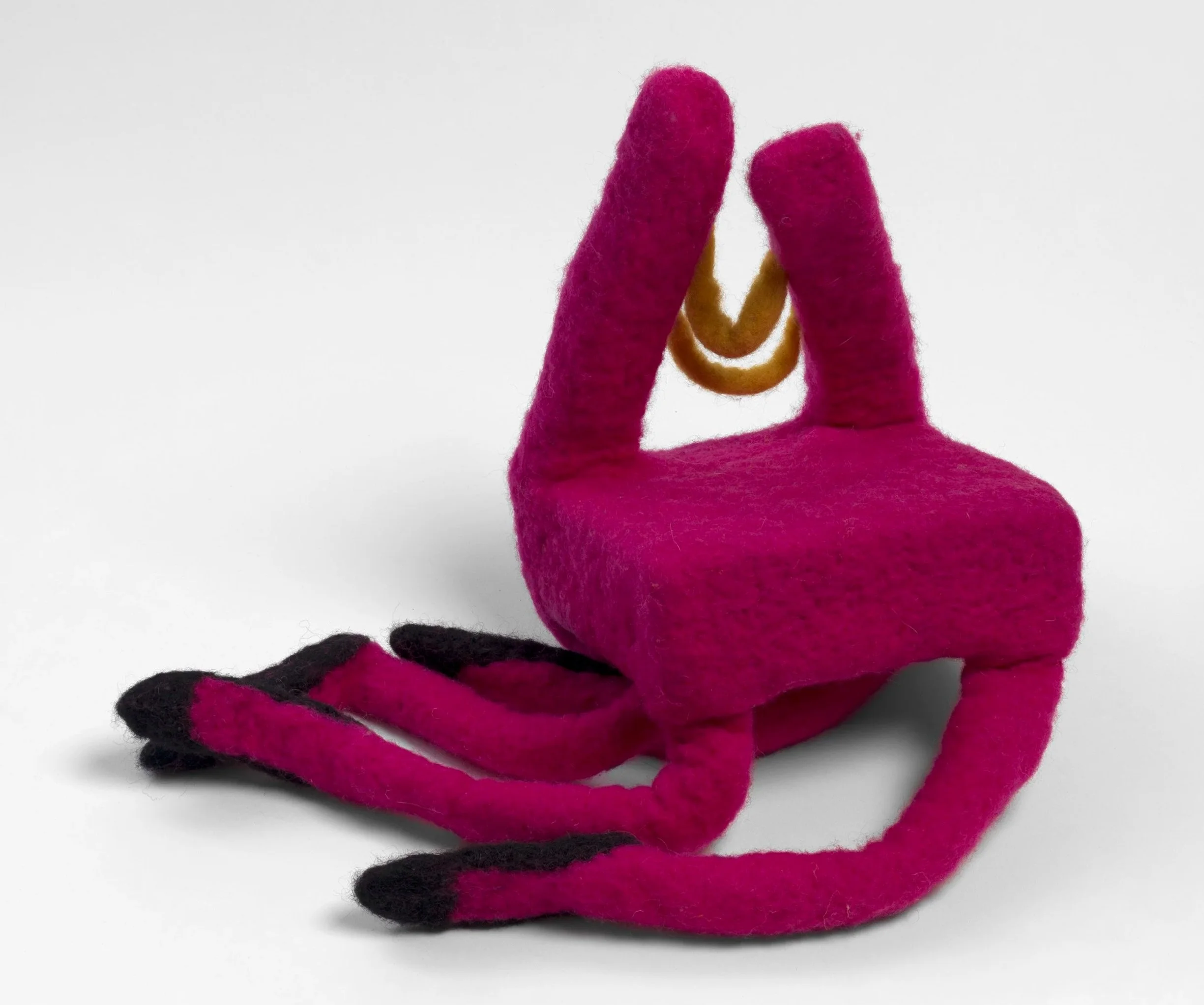 Needle felted chair in hot pink wool, sitting down with soft legs tucked under and to the side. The legs have what looks like black shoes on. The back is made of two vertical pink and two horizontal yellow wool lines..