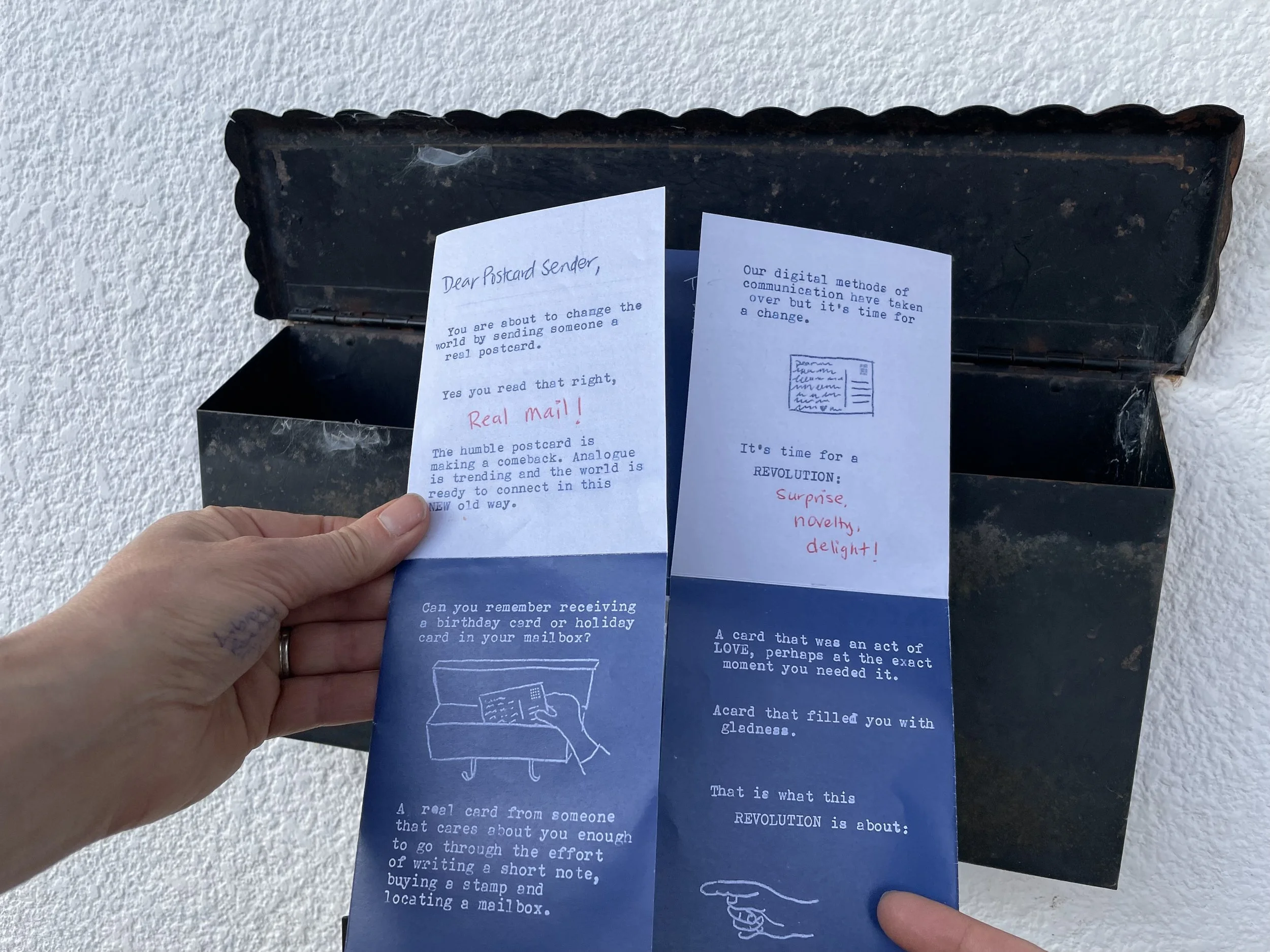 Hands holding a partially unfolded 'One Postcard at a Time: a MANIFESTO! zine in front of mailbox.