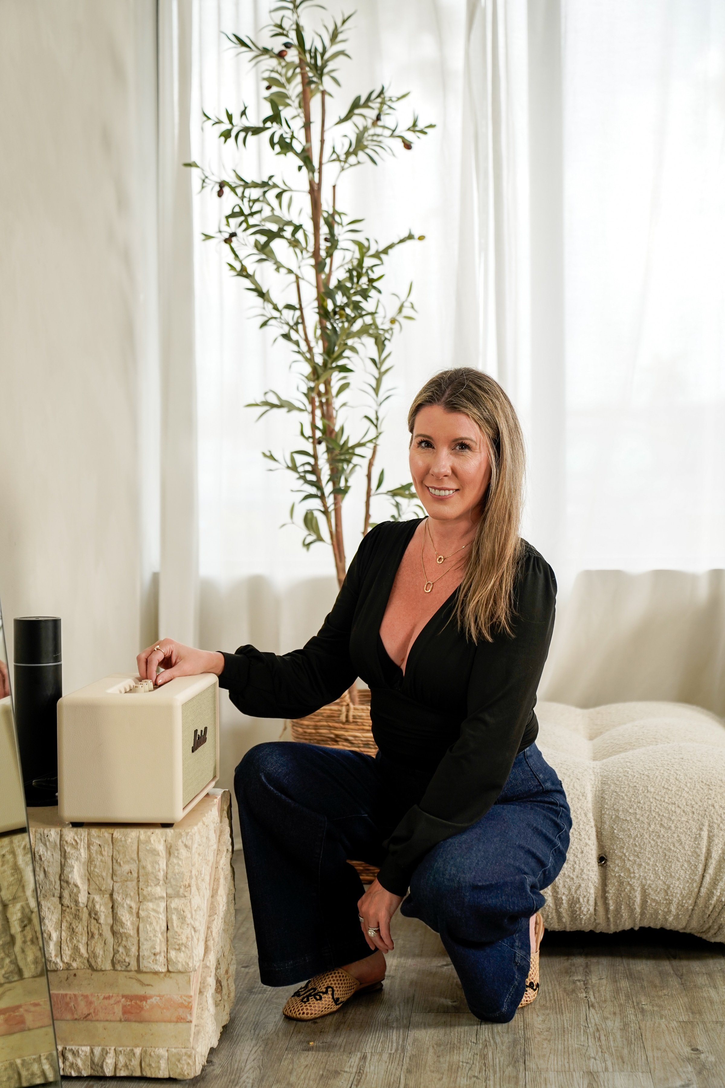 A woman with long blonde hair, wearing a black top and blue jeans, is sitting on her heels in a bright room, adjusting vintage-style speakers on a stone table. Behind her is a large potted plant, a beige couch, and sheer white curtains.
