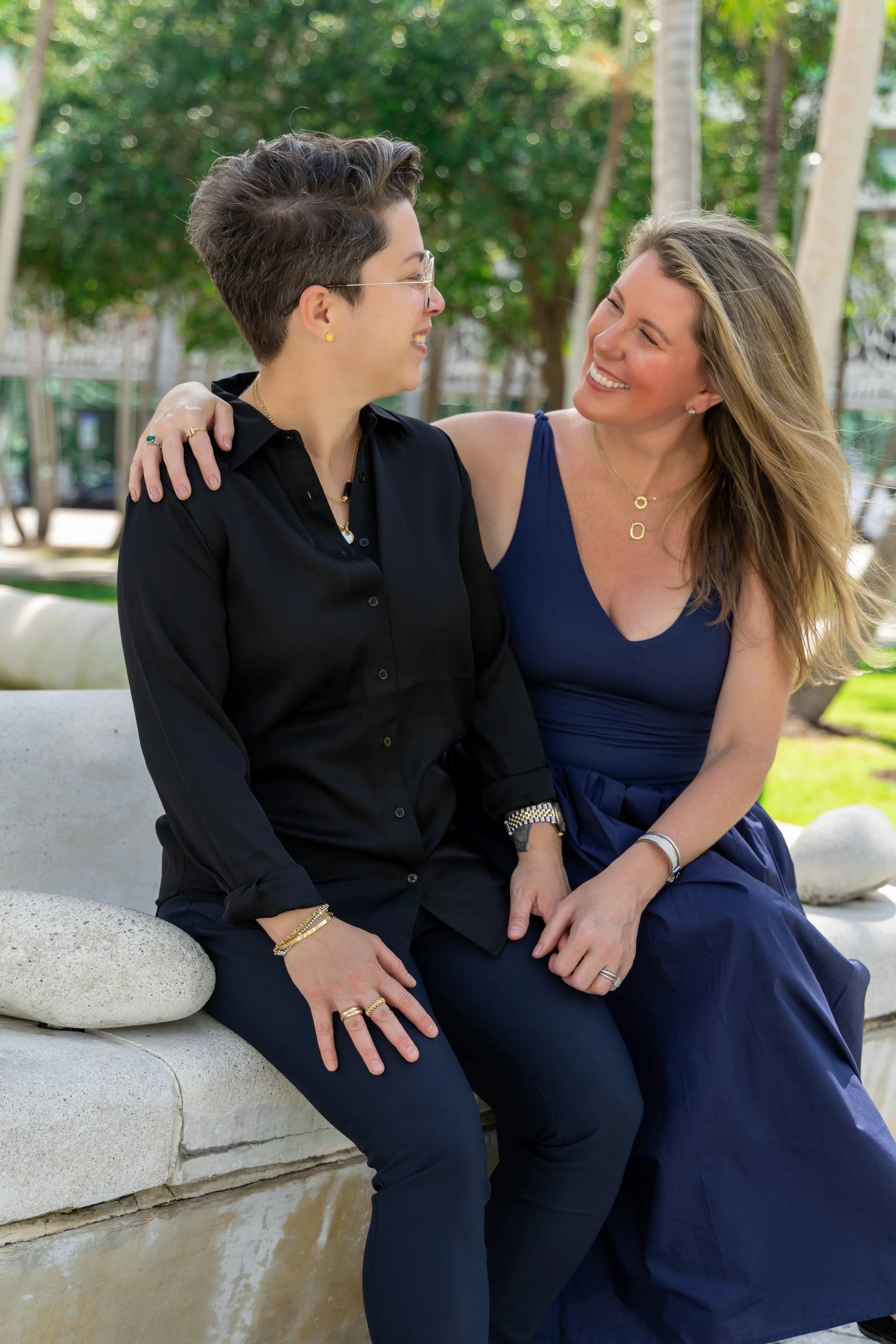 Two women sit close together on a stone bench outdoors, smiling at each other. The woman on the left has short, dark hairstyle, glasses, and is dressed in a black shirt. The woman on the right has long, blonde hair, and is wearing a blue dress. They 