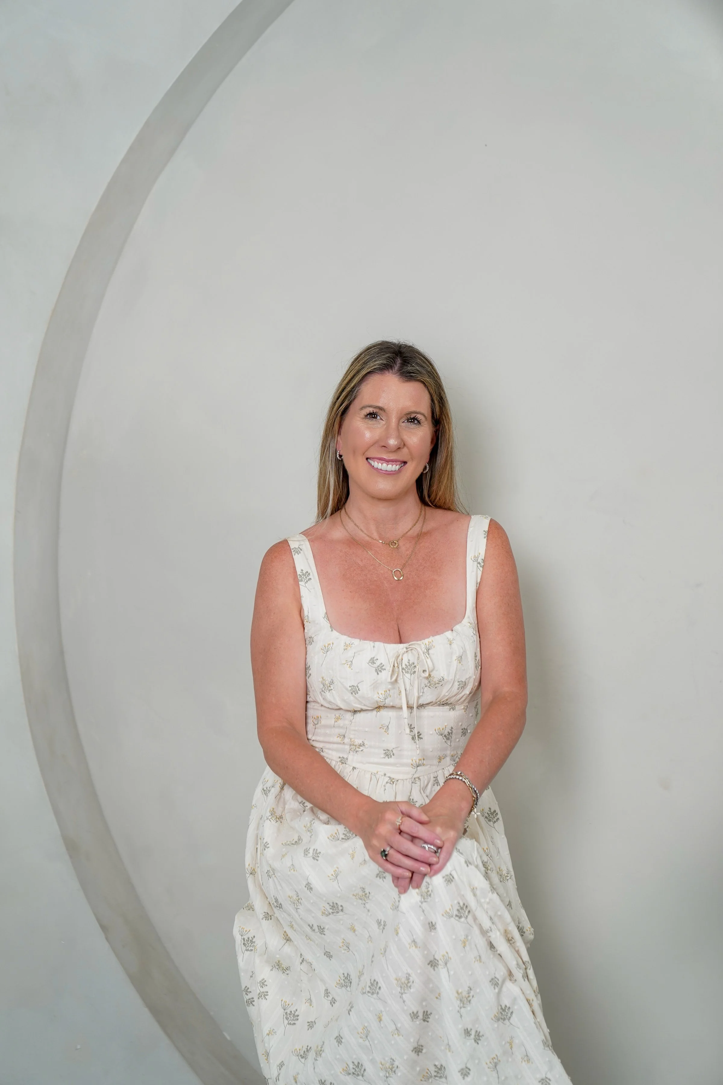 A smiling woman with long blonde hair wearing a white dress with a floral pattern, standing inside a space station or similar structure with a large circular opening.
