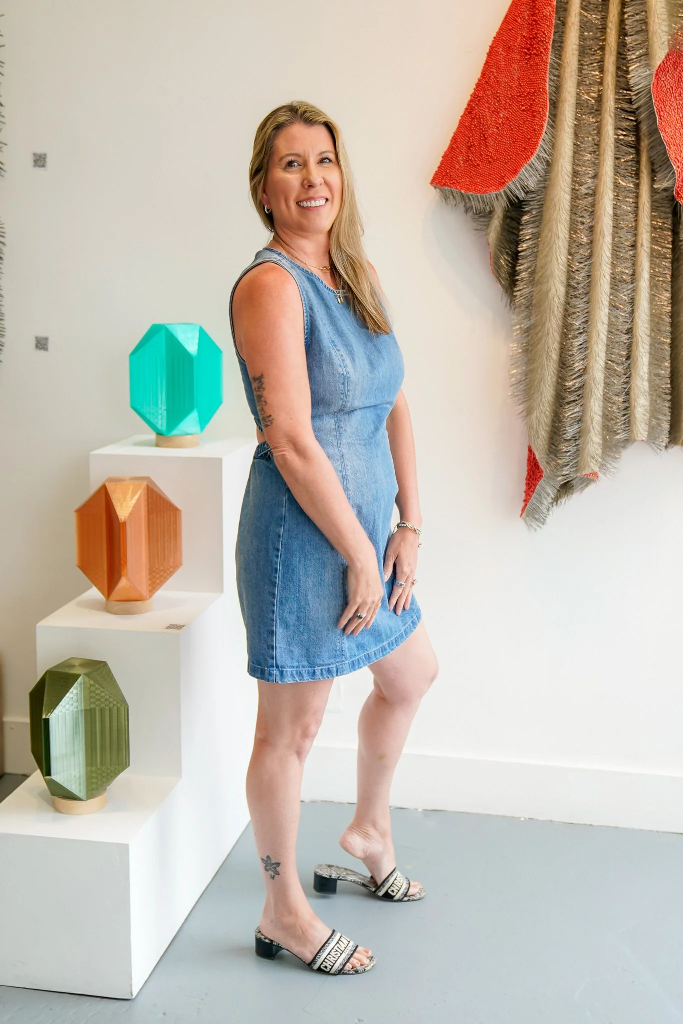 A woman with blonde hair, wearing a sleeveless denim dress and high heel sandals, standing in an art gallery. Behind her are geometric sculptures on a white display and textile wall art on the wall.
