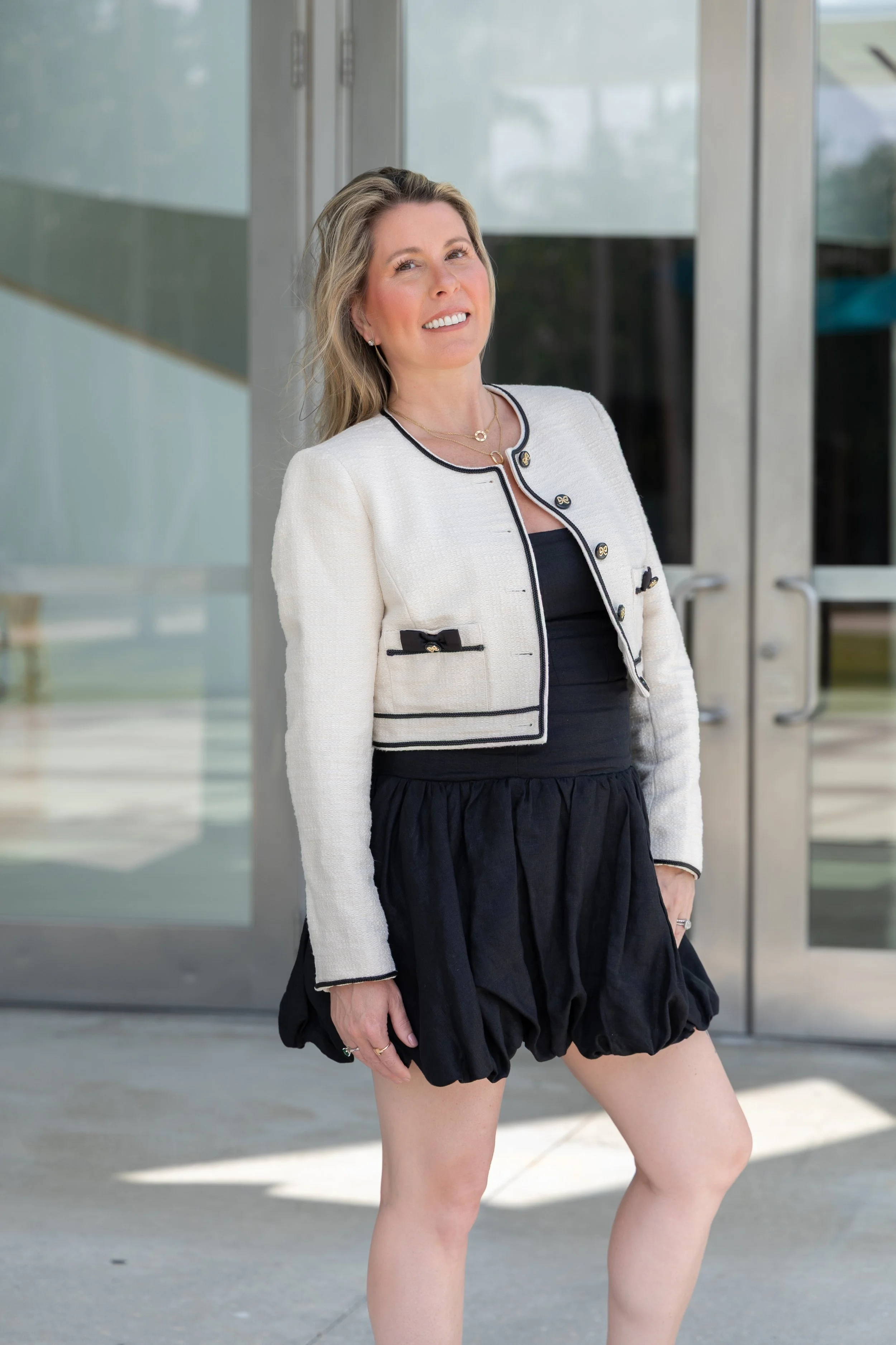 A woman standing outdoors near glass doors, wearing a white cropped jacket with black trim, a black dress, and a black skirt, smiling at the camera.