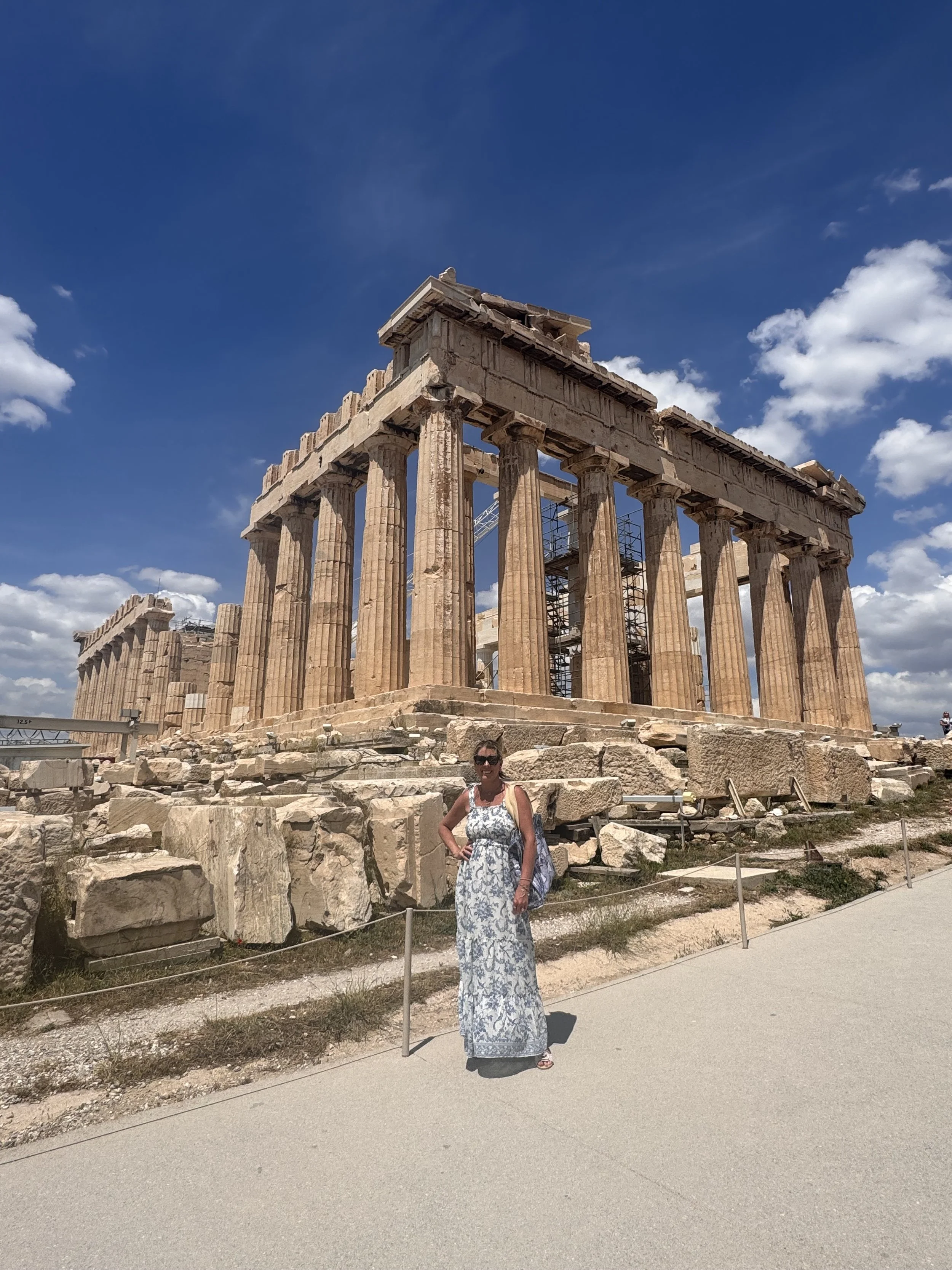 A woman in a long, white and blue patterned dress and sunglasses standing in front of the ancient ruins of the Parthenon on the Acropolis in Athens, Greece, under a partly cloudy blue sky.
