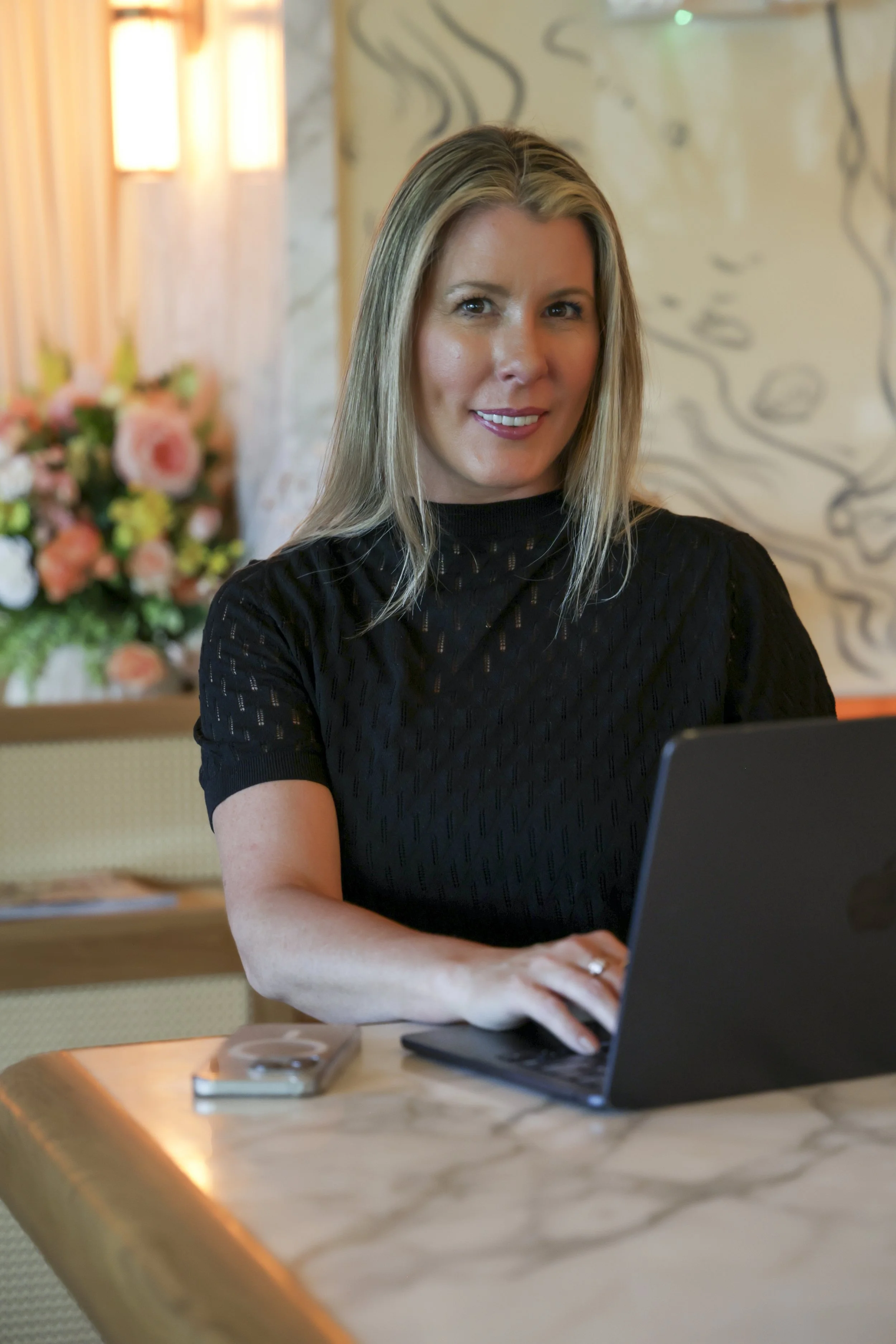 A woman with blonde hair, wearing a black top, working on a laptop at a wooden table in a warmly lit room with floral and abstract artwork on the walls.