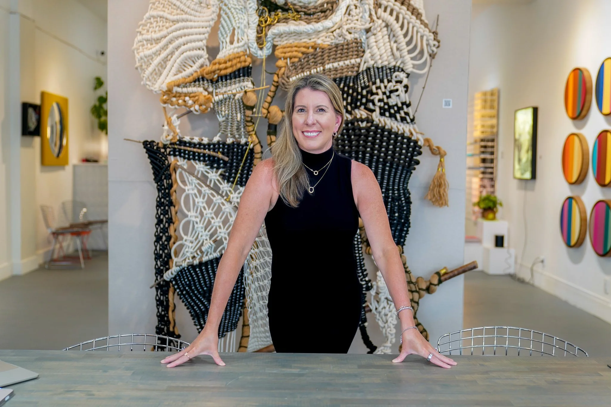 A woman in a black sleeveless top smiling, standing at a table with her hands resting on it, behind her an abstract textile wall art and colorful circular wall decorations are visible in a gallery setting.