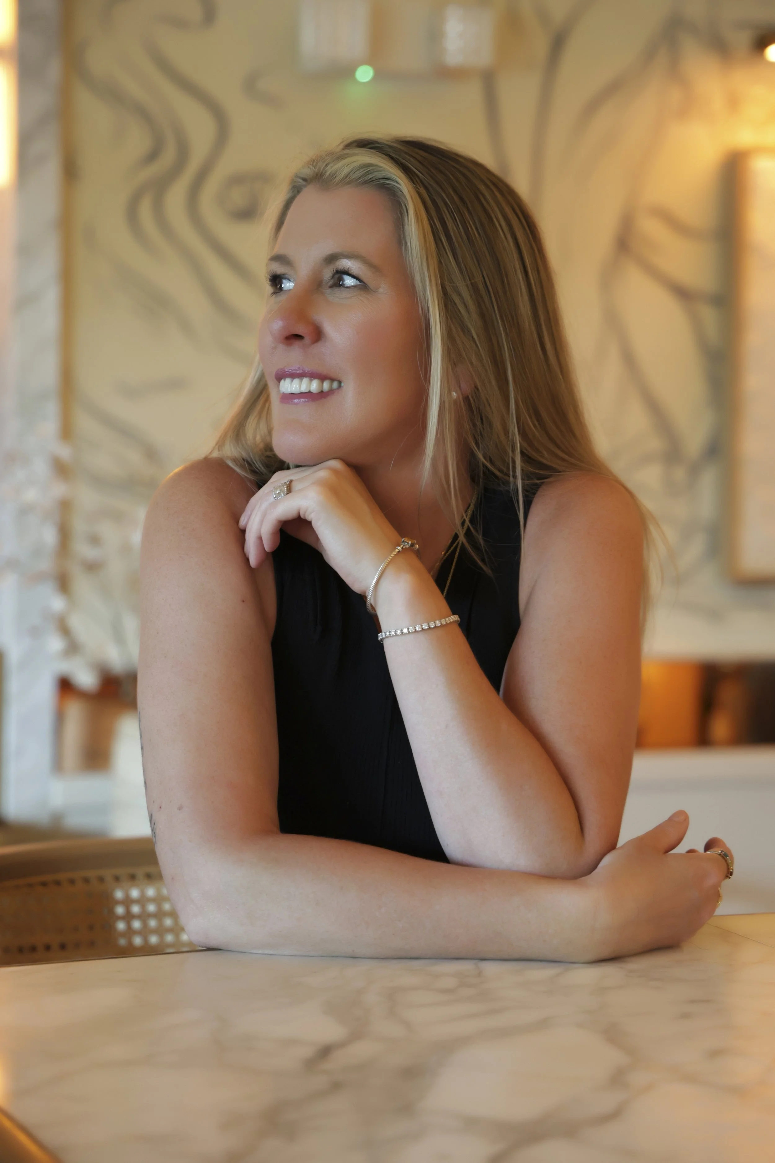 A woman with blonde hair, wearing a black top, sitting at a marble table, smiling and looking to her left, in a warmly lit indoor setting.