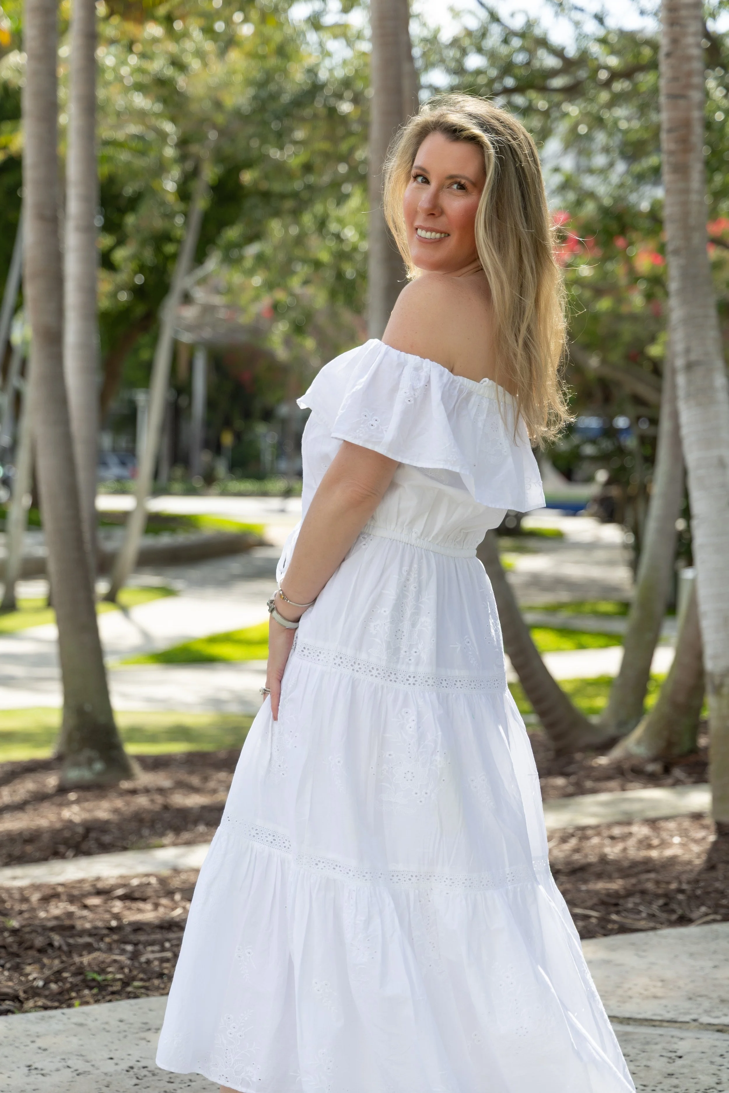 A young woman with blonde hair in a white off-the-shoulder dress smiling outdoors, standing in front of palm trees and greenery.