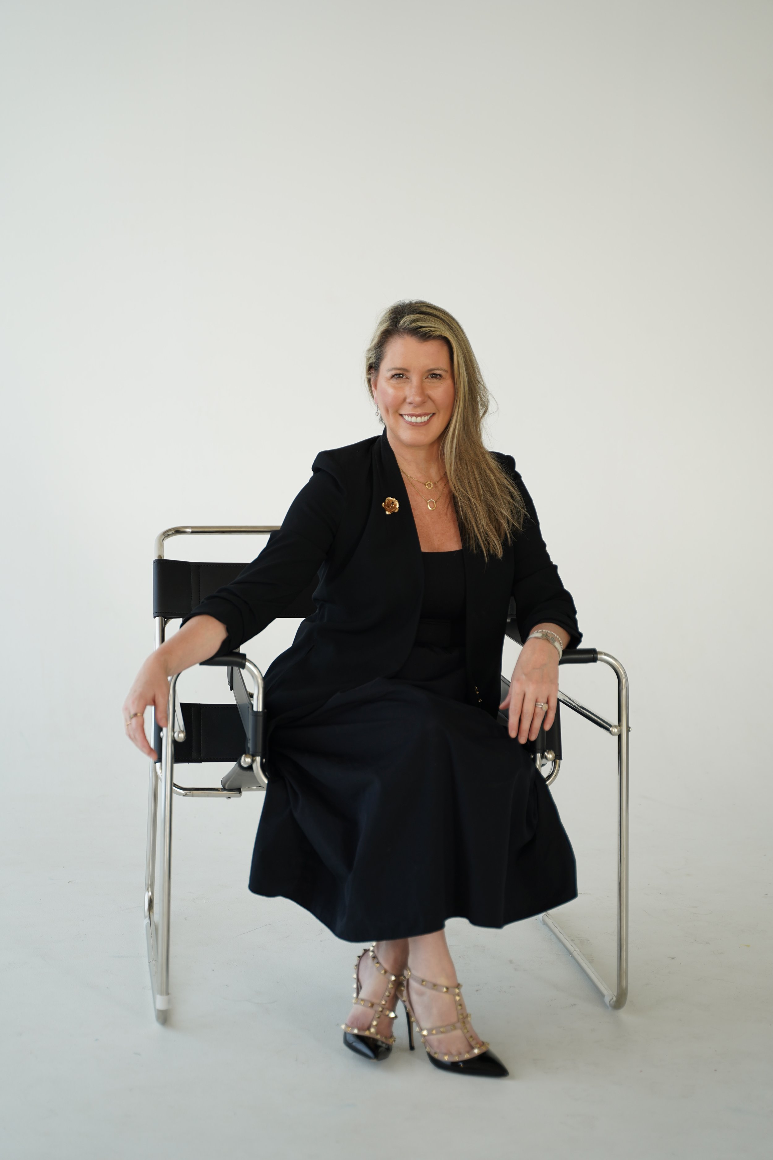A woman with long blonde hair, wearing a black dress and blazer with jewelry, seated in a wheelchair with metal frame, smiling and posing against a plain white background.
