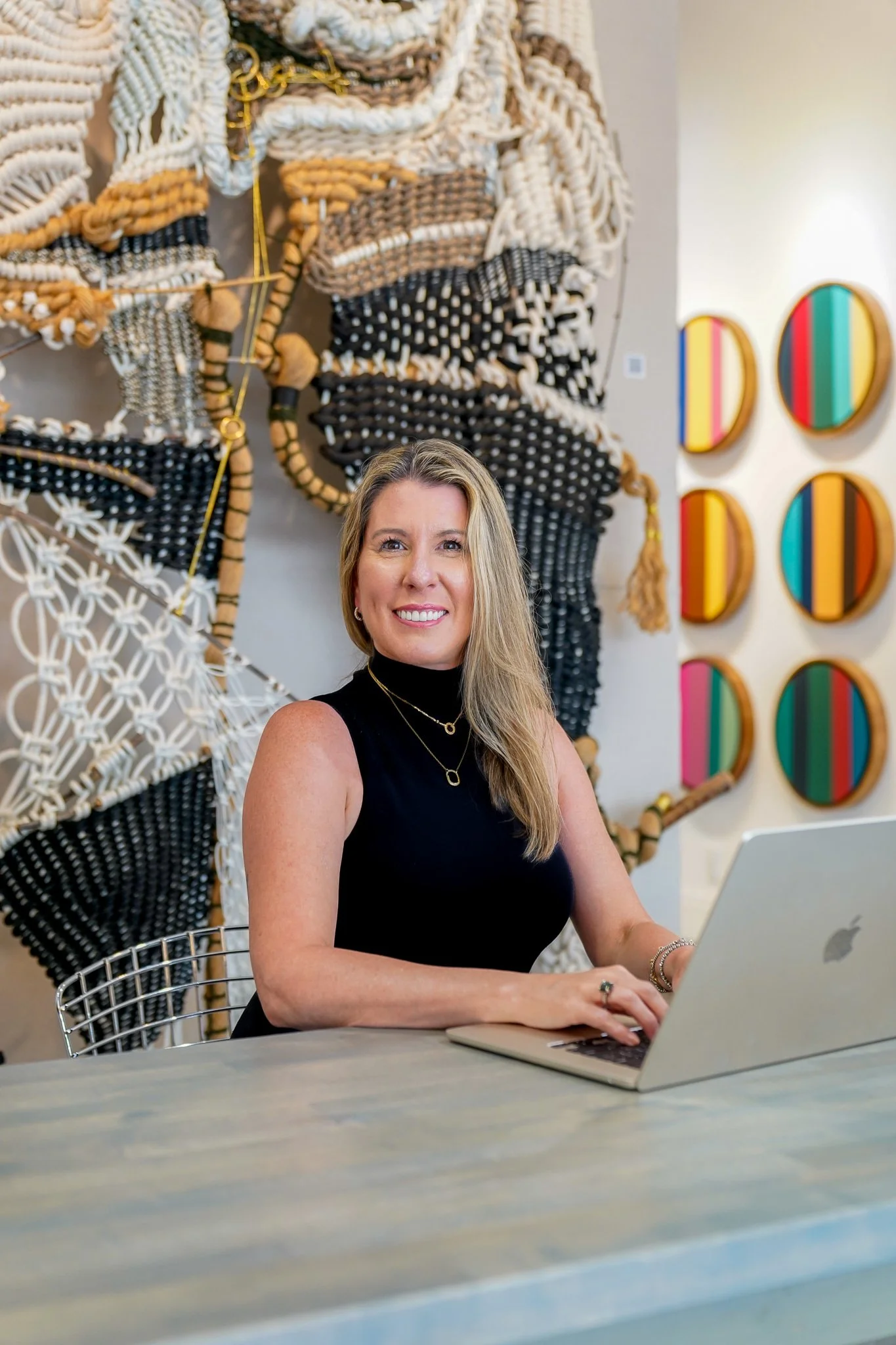 A woman with blonde hair wearing a black sleeveless top, sitting at a desk with a silver laptop, smiling and working in an art gallery or exhibition space with colorful circular wall art and textured woven wall hangings behind her.