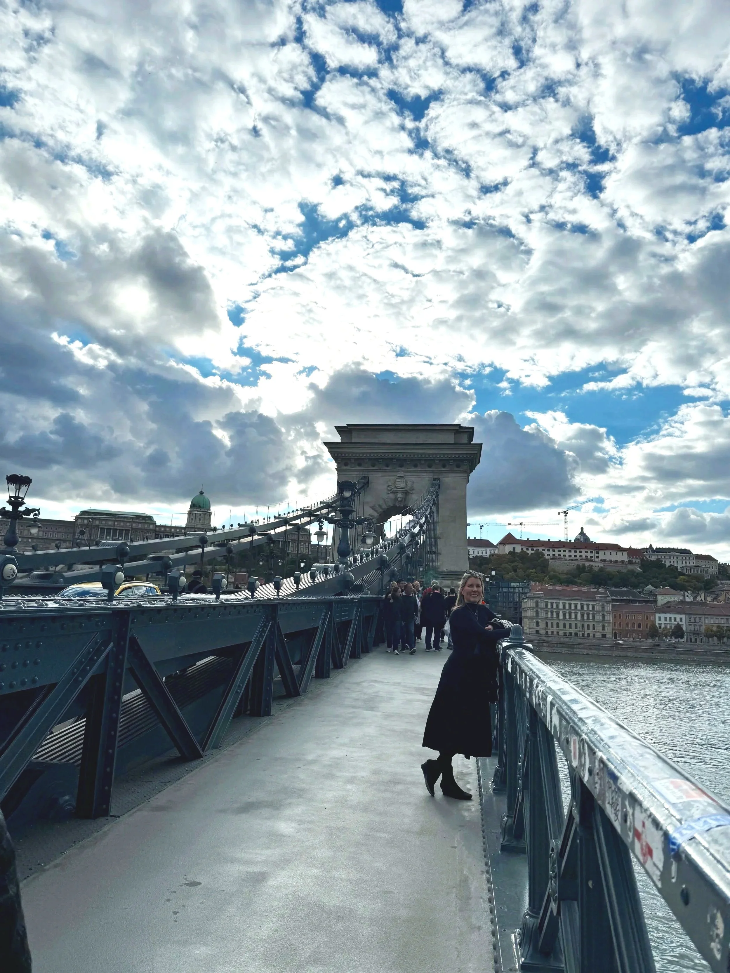 People walking on a bridge over a river with a cityscape and a cloudy sky in the background.