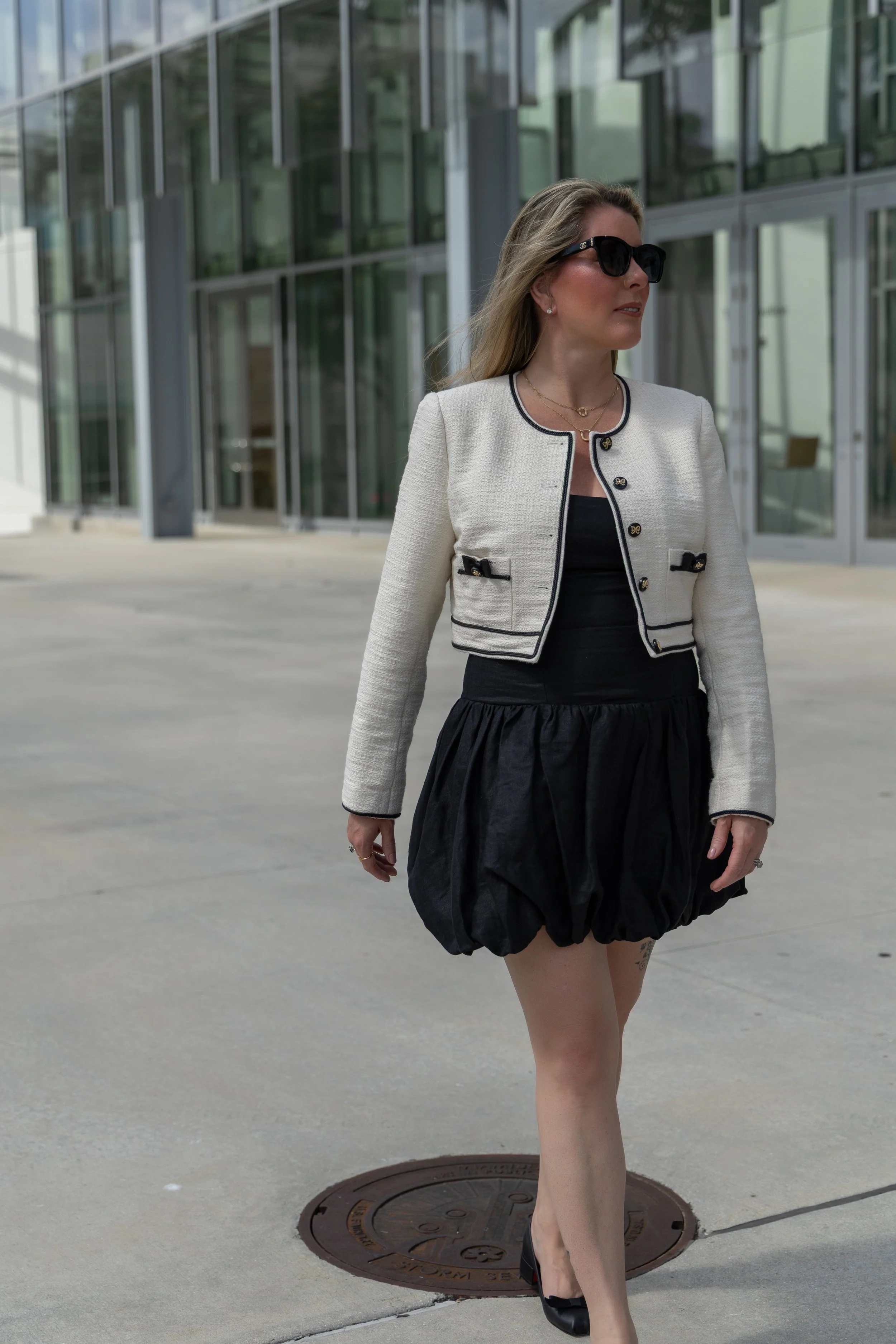 A woman walking outdoors in front of a modern glass building, wearing sunglasses, a white jacket with black trim, a black dress, a ruffled black skirt, and black high heels.