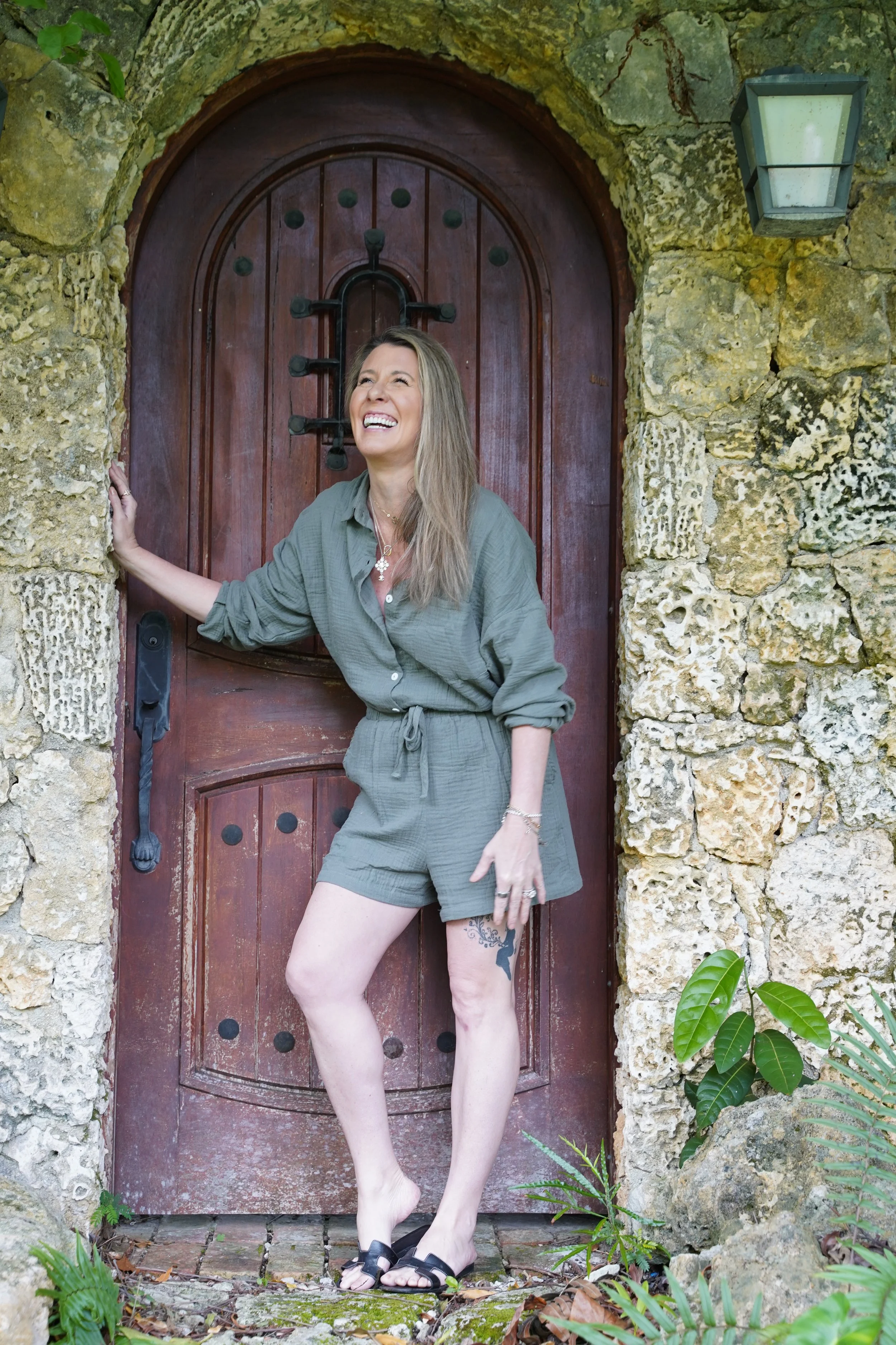 A woman with long blonde hair, wearing a green jumpsuit and black sandals, posing cheerfully in front of a wooden door set in a stone wall surrounded by greenery.