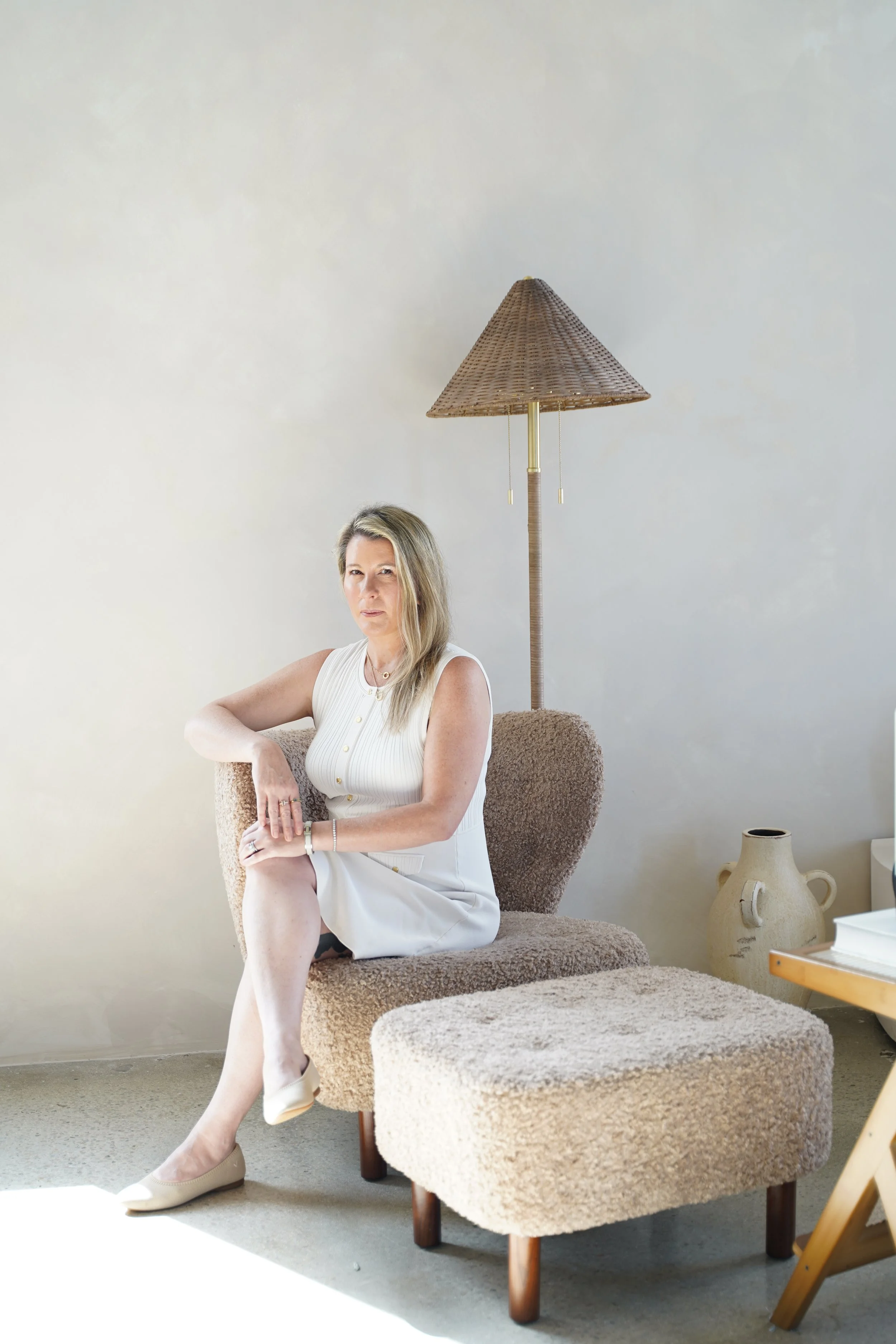 A mature woman with blonde hair sitting on a plush, beige armchair in a minimalist room. She wears a sleeveless white dress and beige heels. An antique ceramic jug and a bedside table with books are visible in the background, with a tall wicker floor lamp behind her.