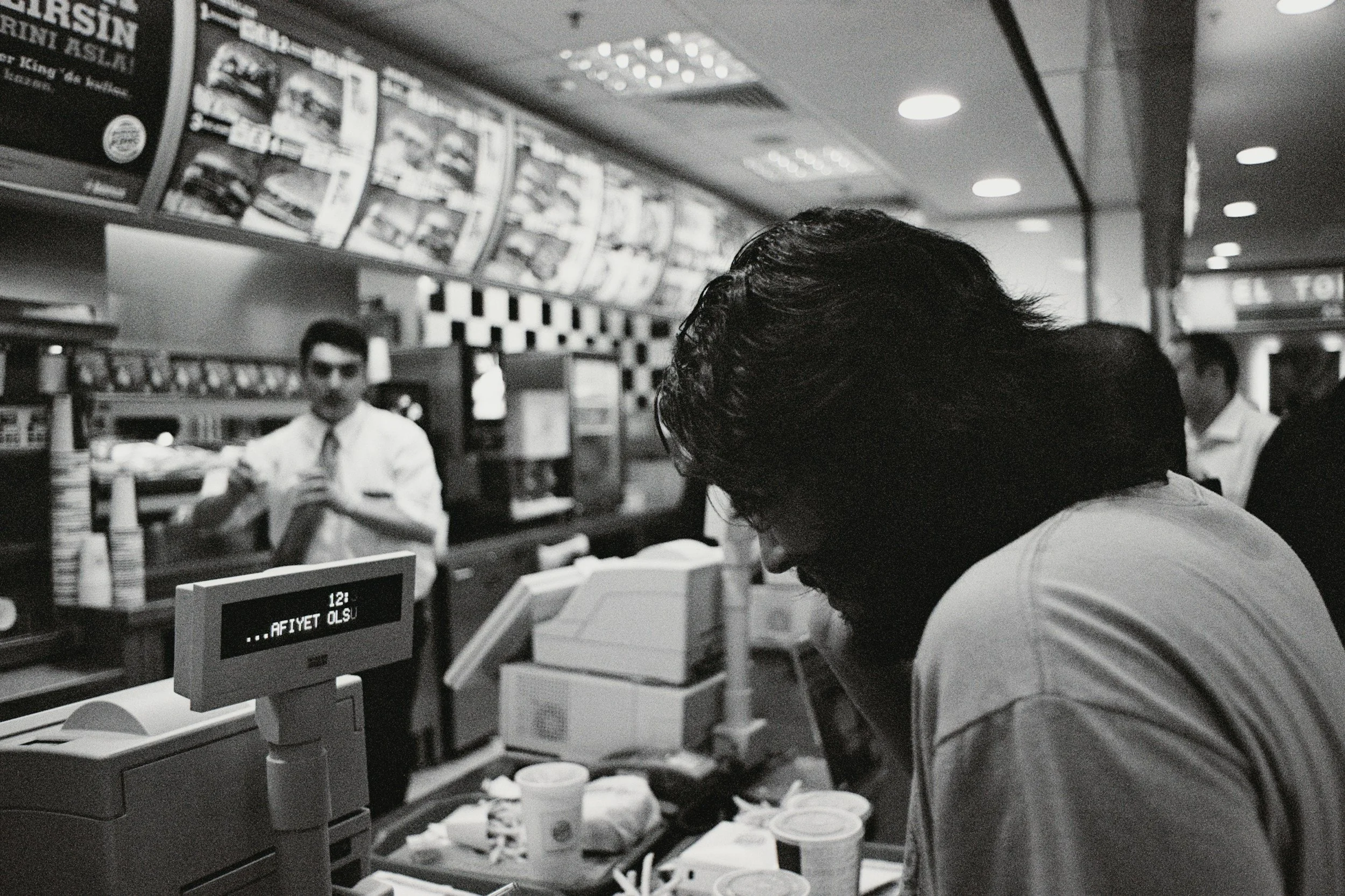 Black and white photo of a man leaning over the counter at a fast food restaurant, showing a busy counter with food containers, a cash register, a digital display, and a worker in the background preparing food.