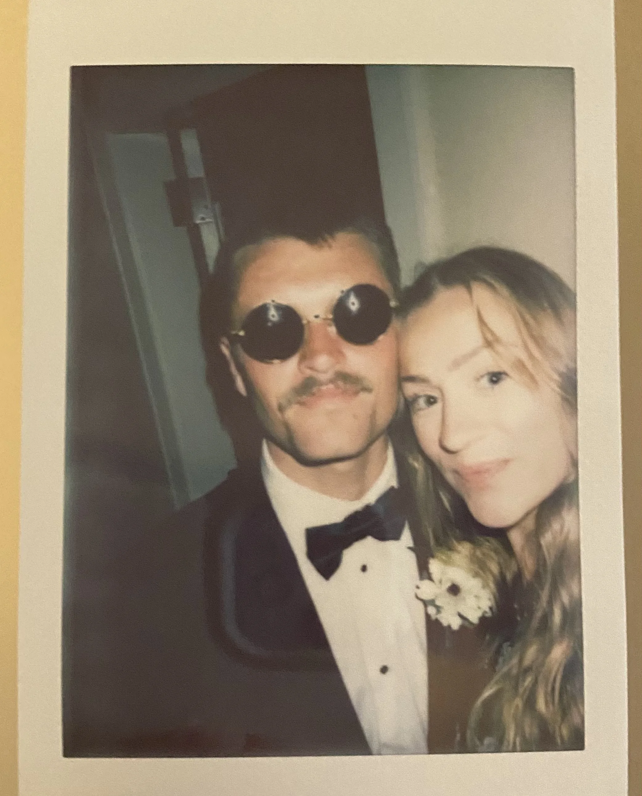 A man in a tuxedo and sunglasses standing next to a woman with long hair at a formal event.