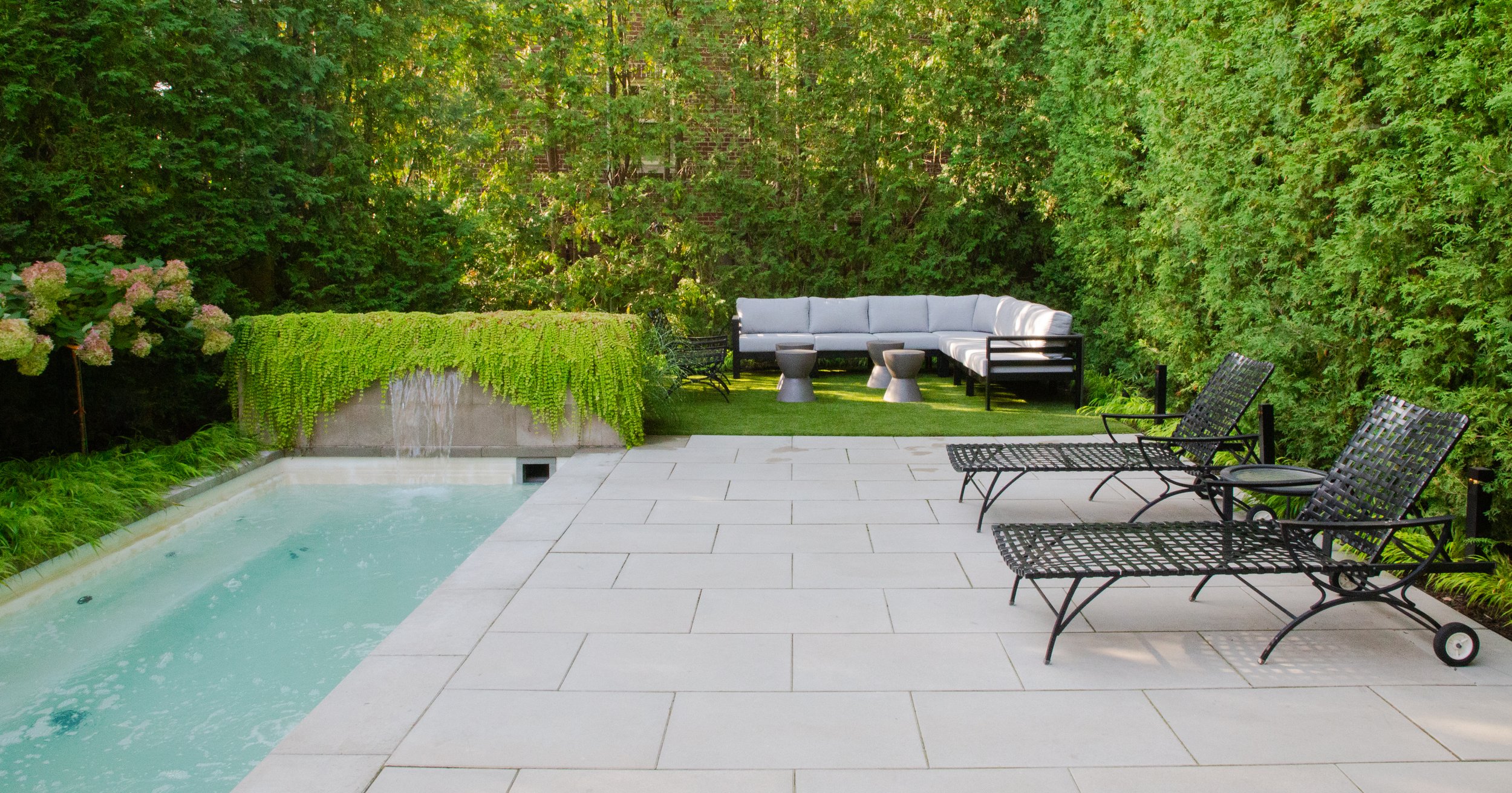 A backyard patio with a swimming pool, black metal lounge chairs, a white corner sofa, small tables, lush green trees, and flowering plants.