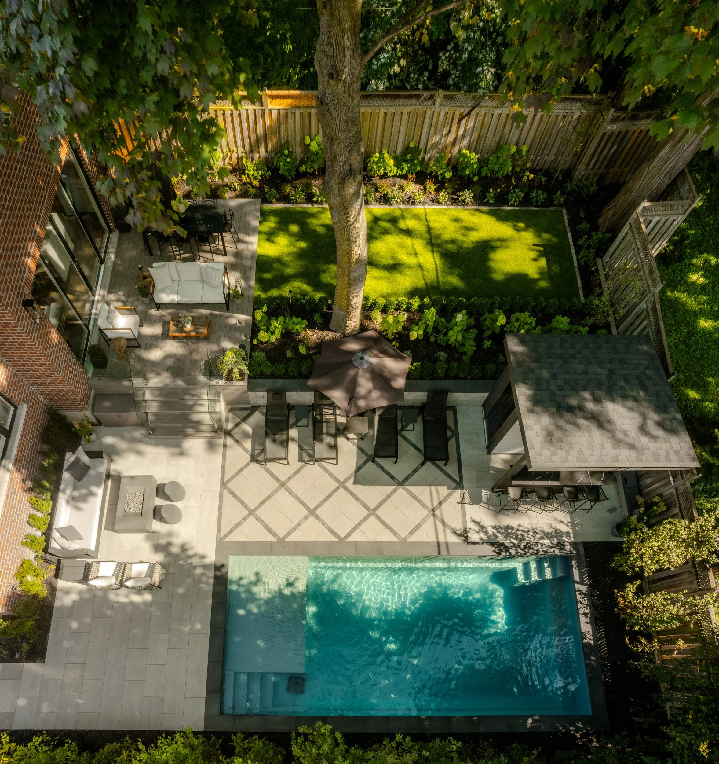 A view of a backyard with a swimming pool, a patio with outdoor furniture, a yard with grass, trees, and a wooden fence.
