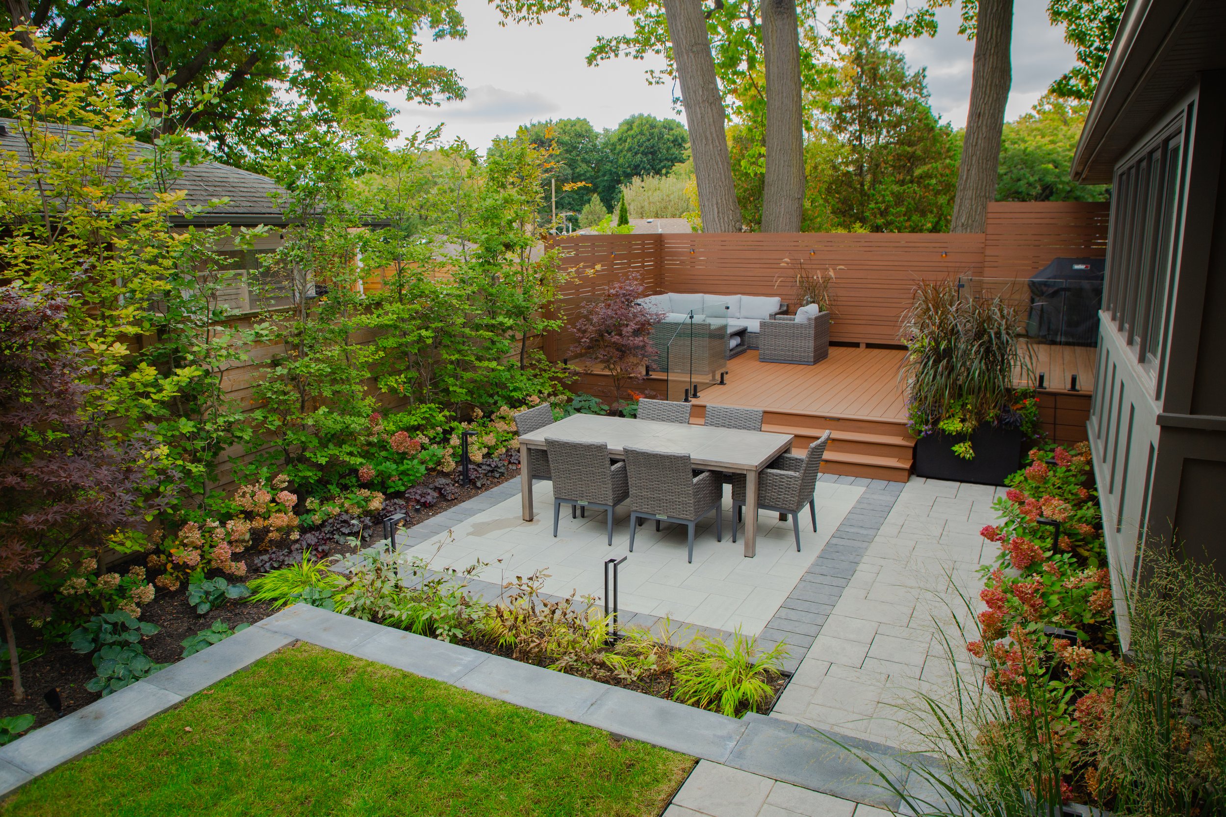 A spacious backyard patio with a dining area with a table and chairs, a wooden deck with outdoor furniture, lush green trees and plants, and a privacy fence, during daytime with overcast sky.