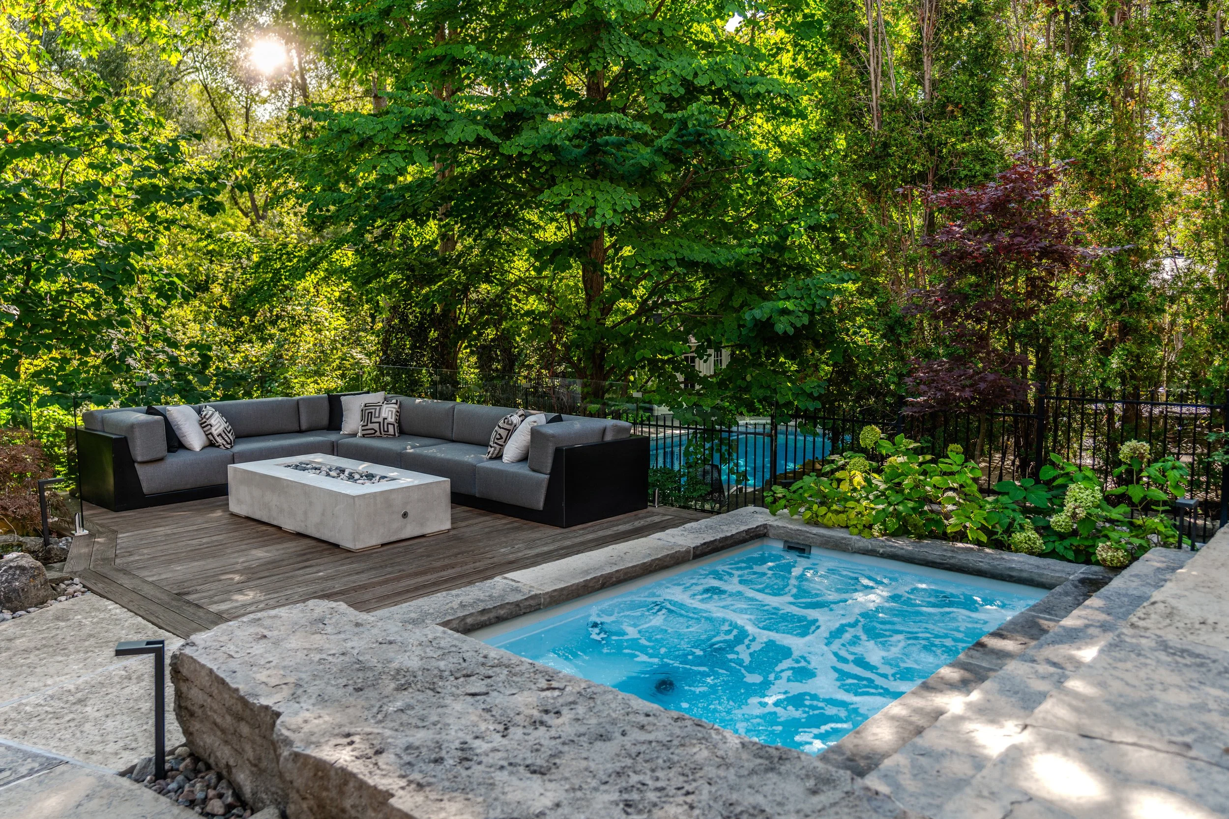 Outdoor patio area with gray sectional sofa with patterned pillows, stone fire pit, and pool, surrounded by lush green trees and bushes with sunlight filtering through.