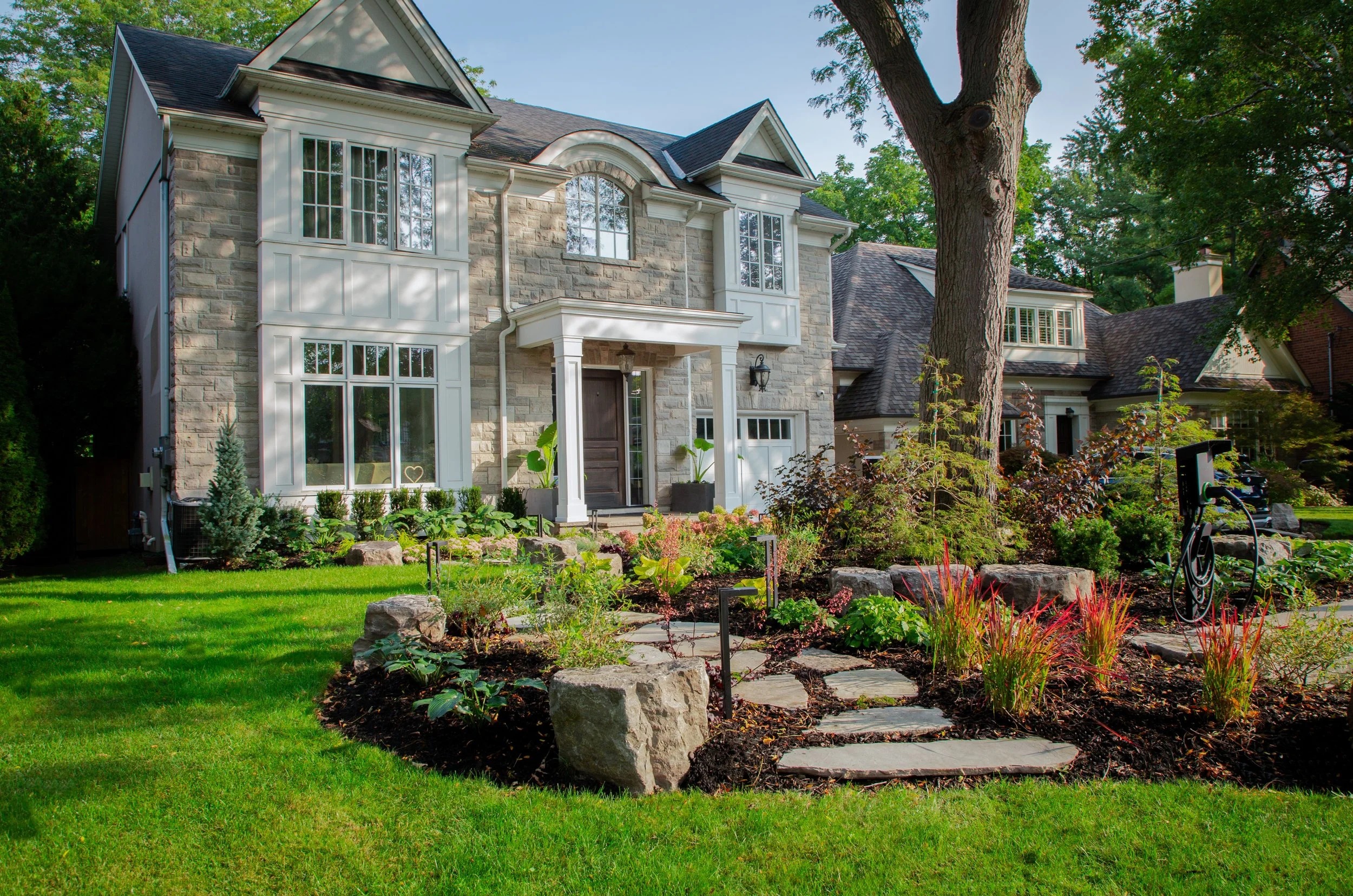 Front view of a house with stone and white exterior, large windows, and a landscaped garden with a stone pathway and lush green lawn.