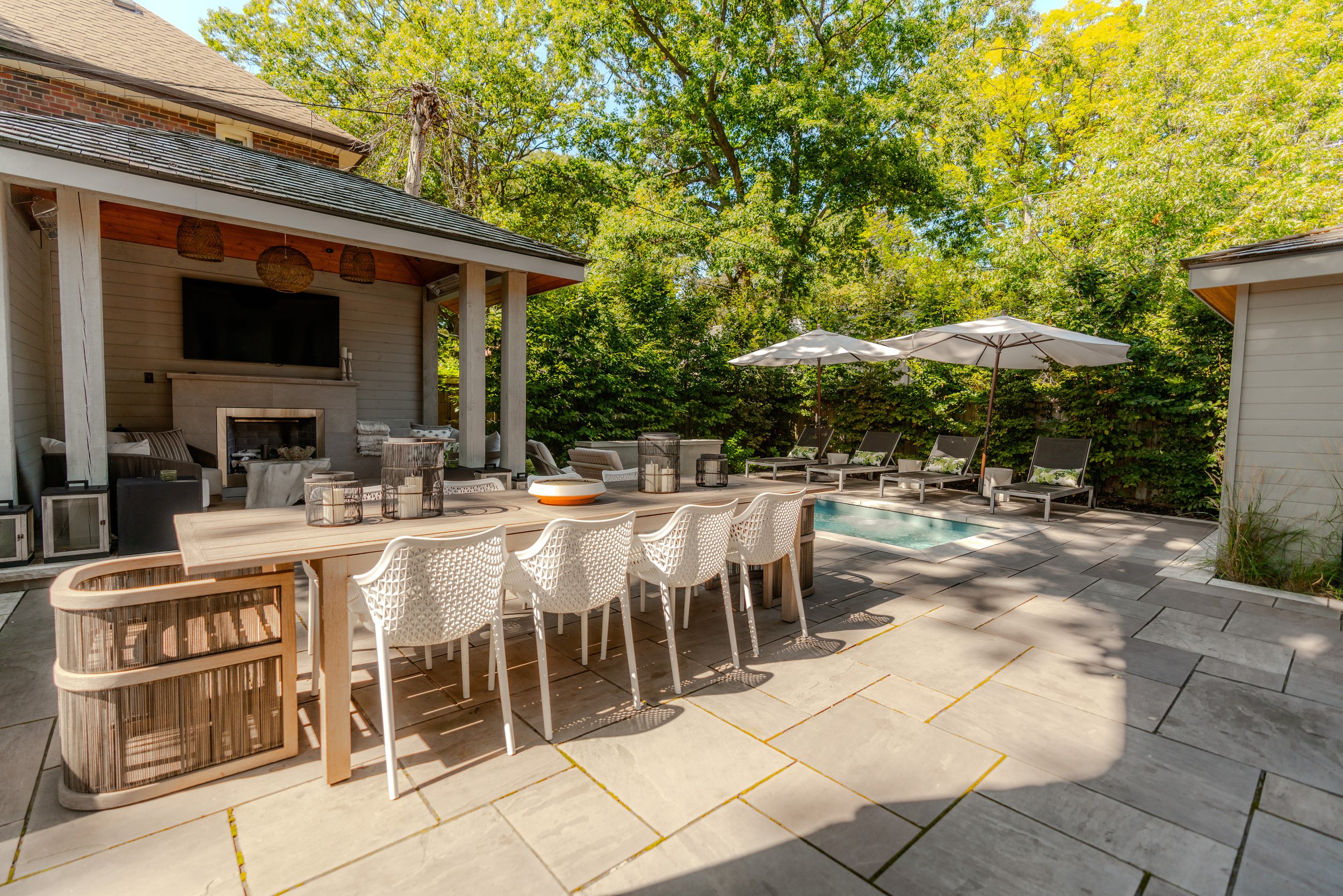Beautiful backyard with outdoor dining area, pool, and lounge chairs under umbrellas, surrounded by lush green trees.