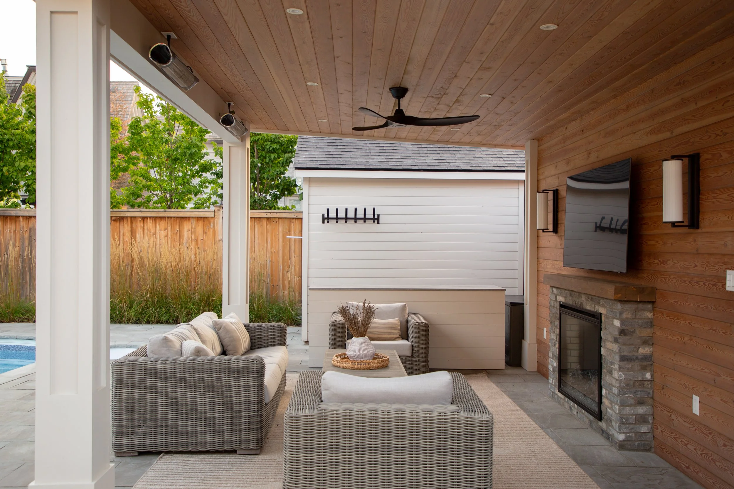 Covered outdoor patio with wicker furniture, a fireplace, a wall-mounted TV, and a ceiling fan. The patio has wood-paneled ceilings and is adjacent to a yard with a wooden fence and green trees.
