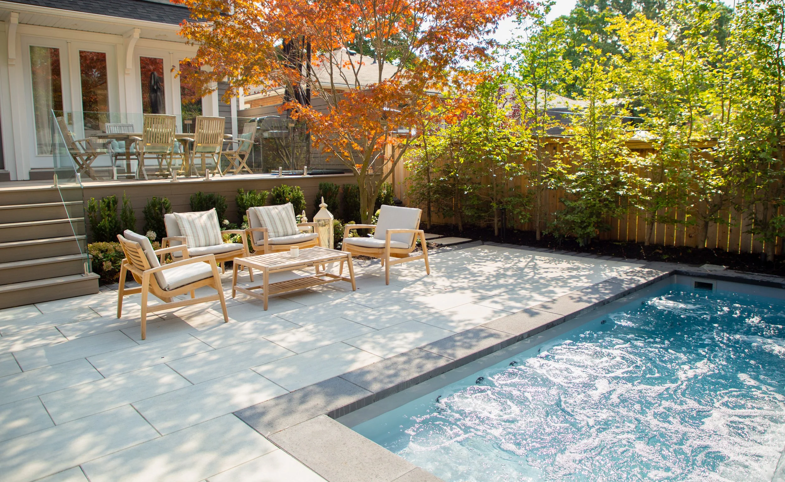 A backyard patio with outdoor furniture, including chairs and a table, next to a swimming pool, surrounded by trees with green and orange leaves.