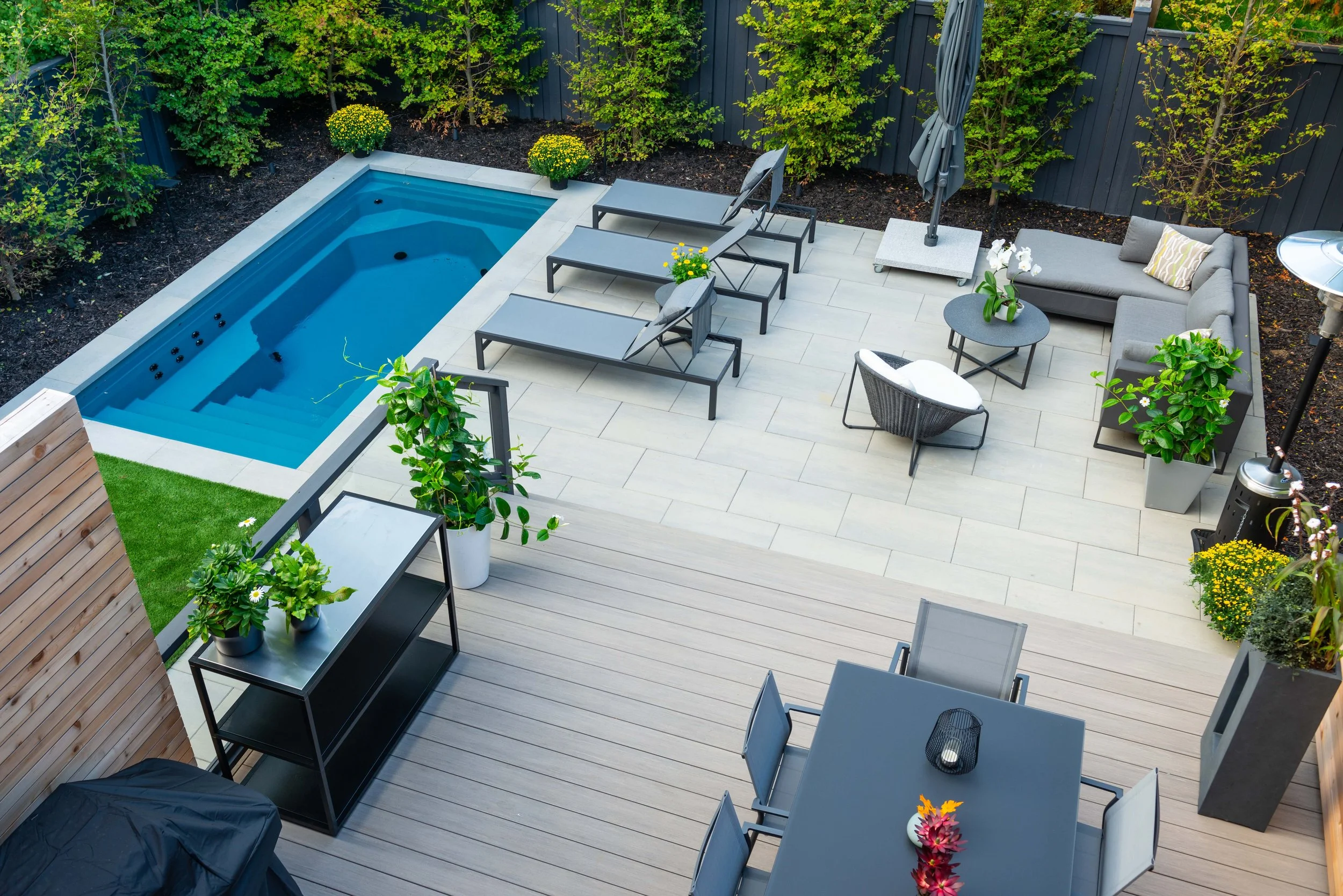 A modern backyard patio with a small pool, lounge chairs, a seating area with a sofa and coffee table, potted plants, and an umbrella, surrounded by greenery and a gray fence.