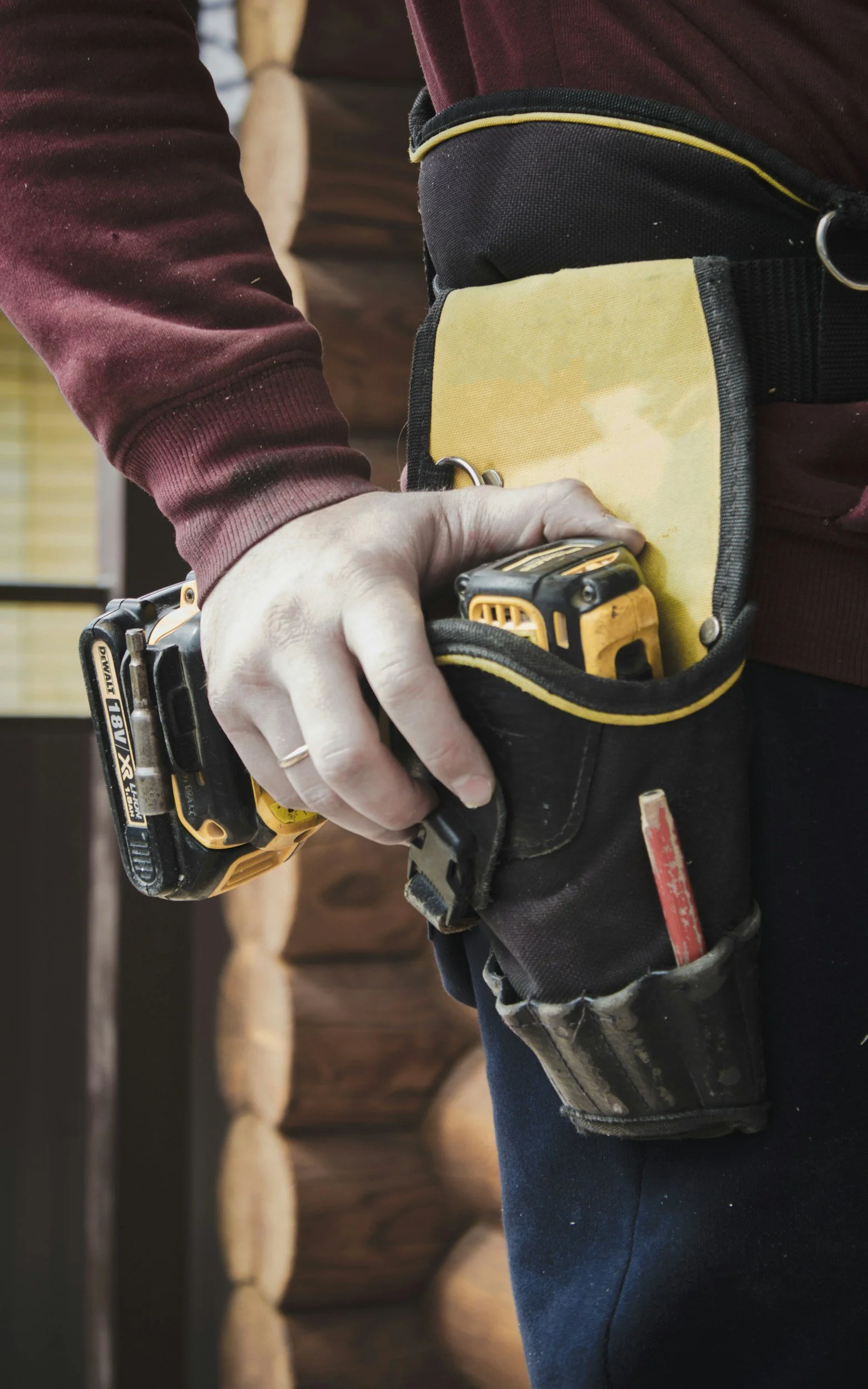 Close-up of a person in a tool belt holding a cordless drill, with a wood wall in the background.