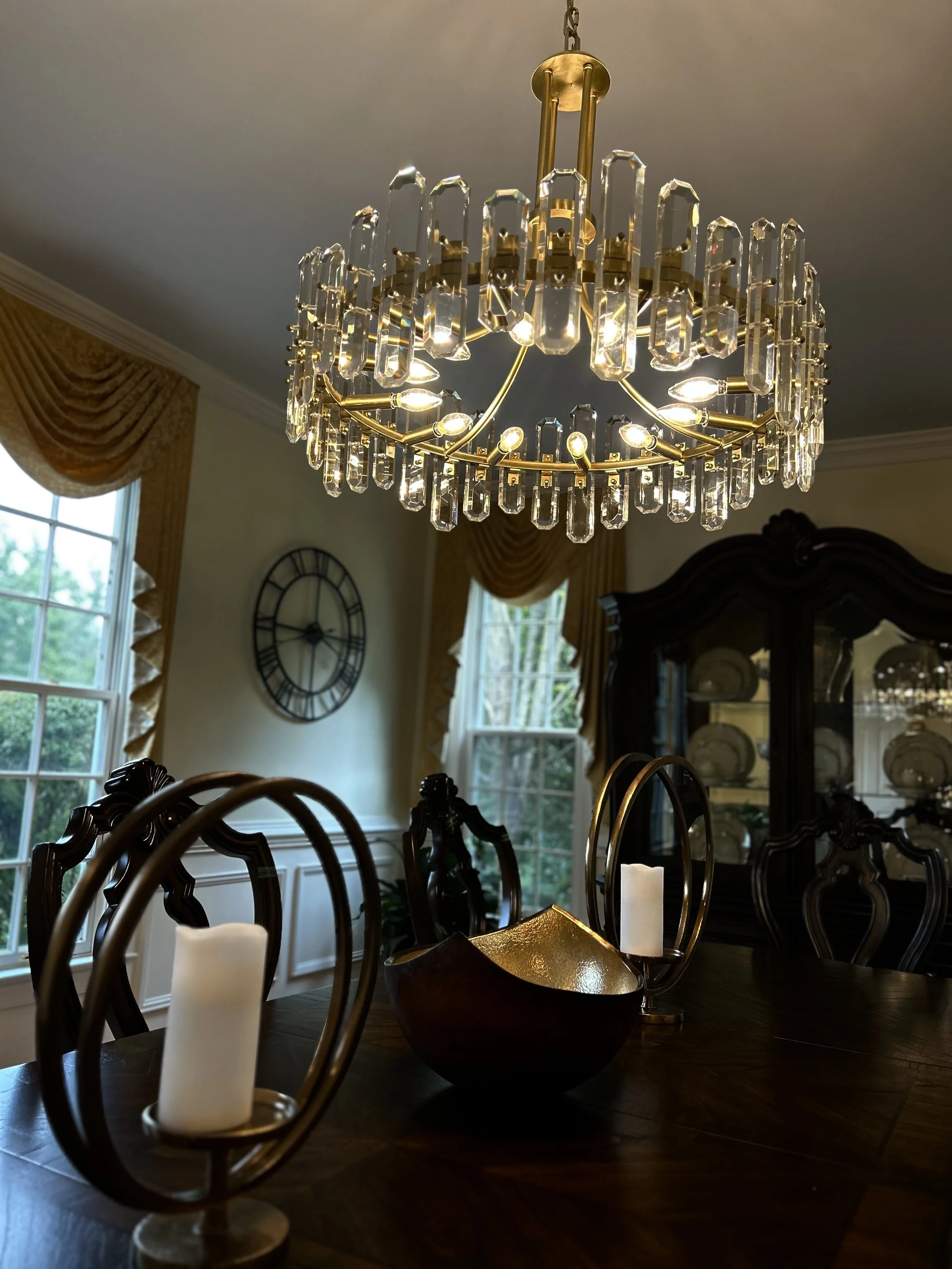 A chandelier with crystal accents hanging over a wooden dining table in a well-lit dining room. The table features decorative candle holders and a bowl, surrounded by dark wooden chairs. Large windows with gold curtains and a wall clock are visible in the background.