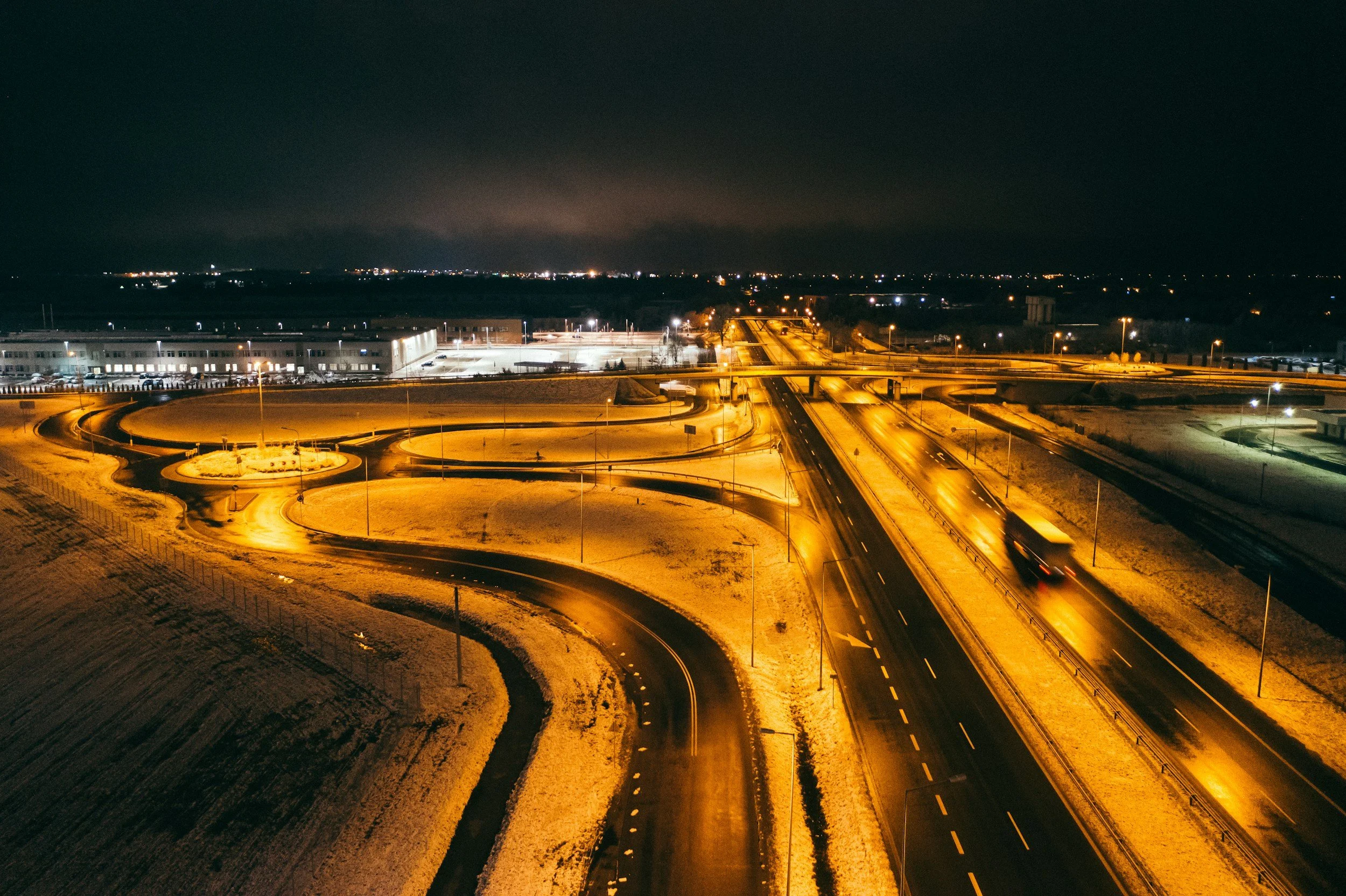 Aerial view of a highway and roundabouts at night with streetlights; snow-covered landscape with buildings in the background.