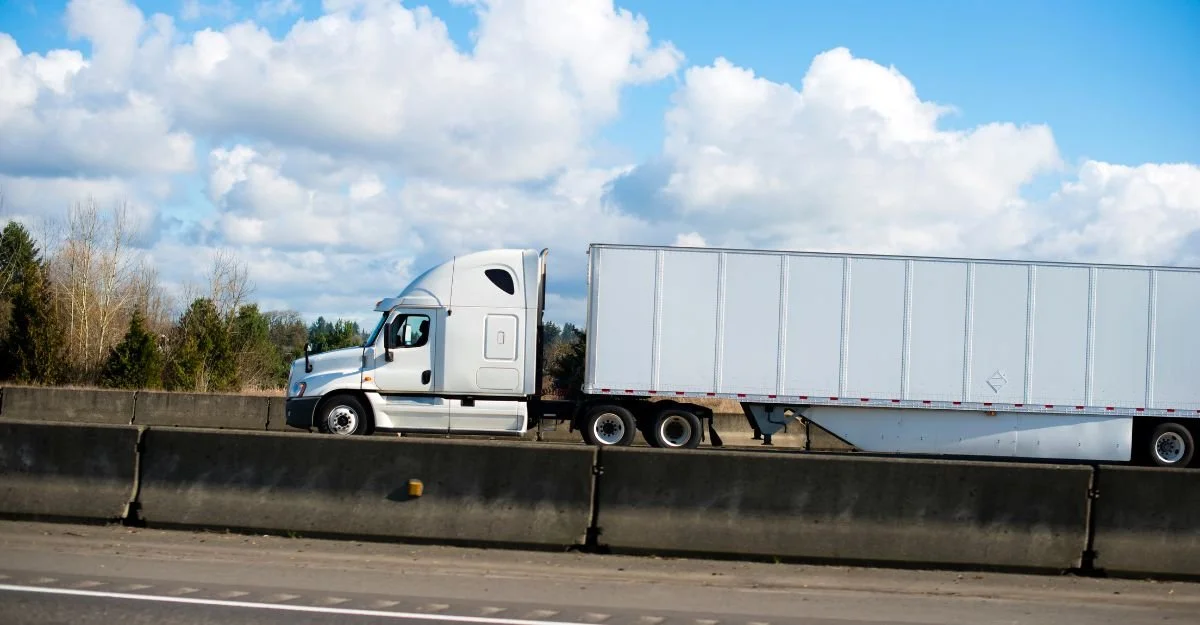 White semi-truck with trailer on highway, concrete barrier in foreground, cloudy sky and trees in background.