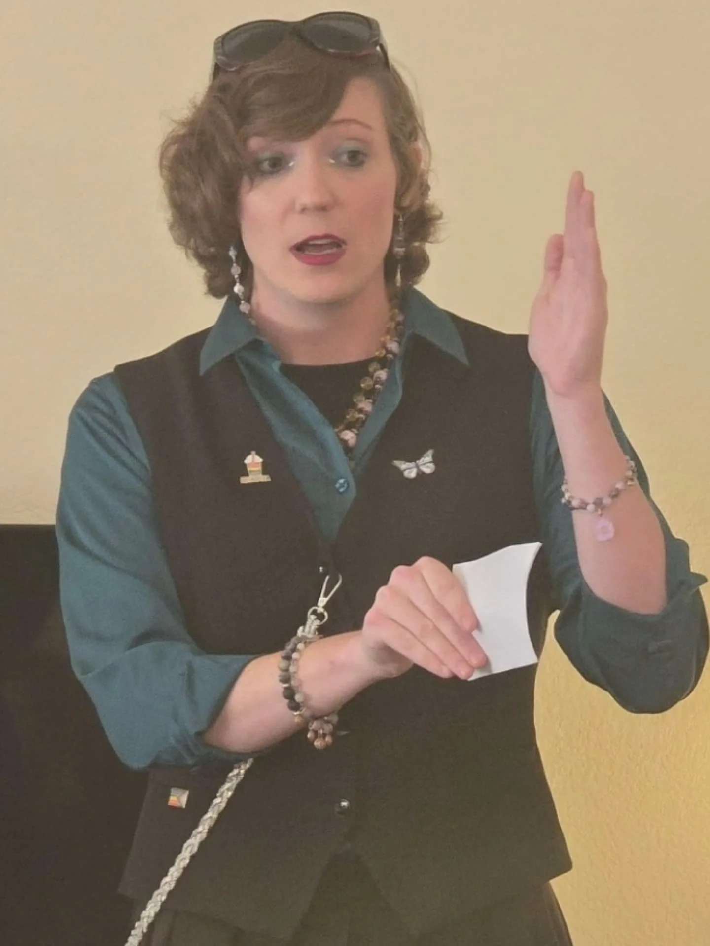 A woman with curly brown hair wearing glasses on her head, earrings, a necklace, and a dark vest over a teal shirt, holding a piece of paper, with her right hand raised and palm facing outward.