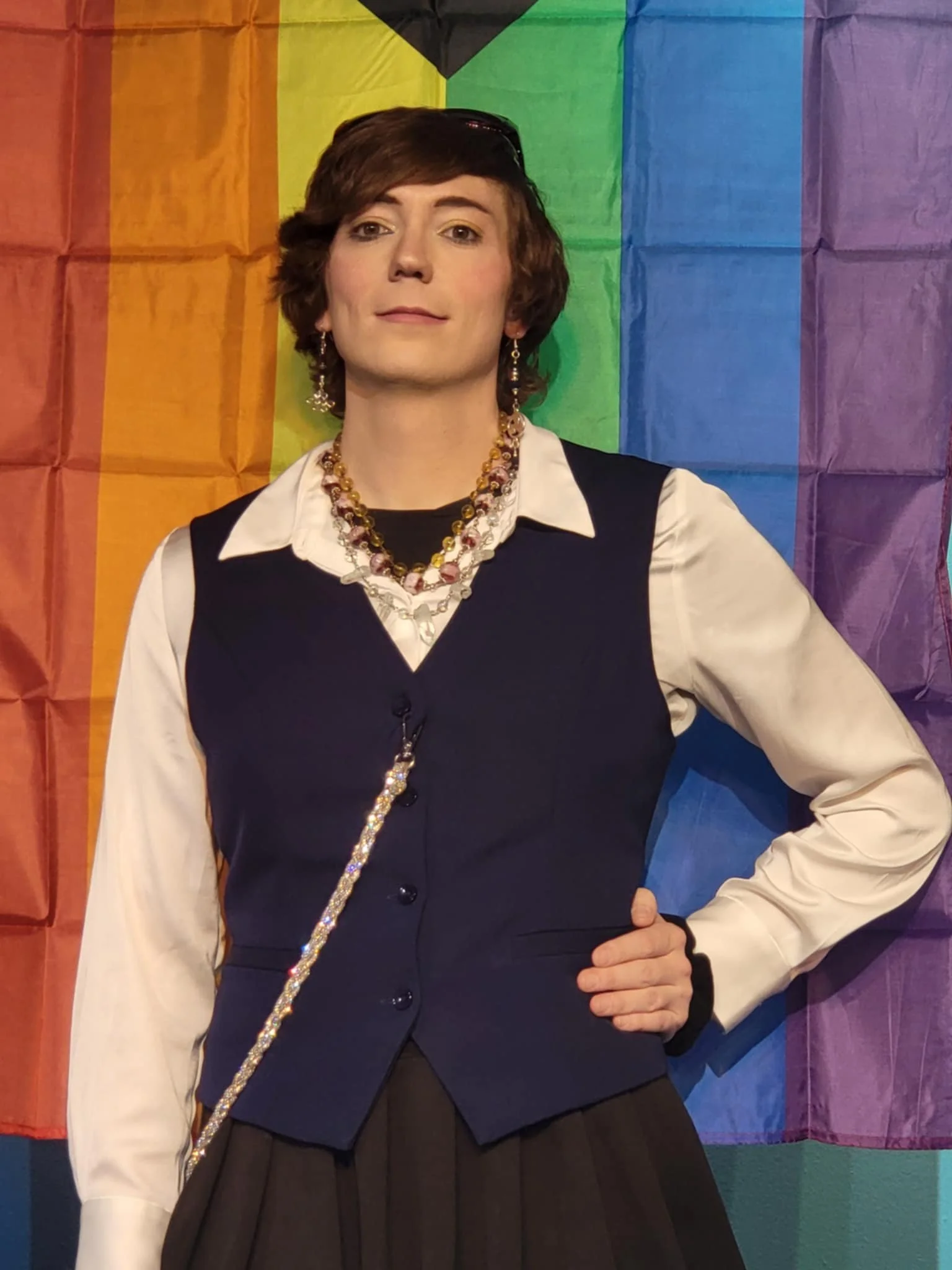 A person standing in front of a rainbow pride flag, wearing a white shirt with a dark vest, multiple necklaces, and earrings.