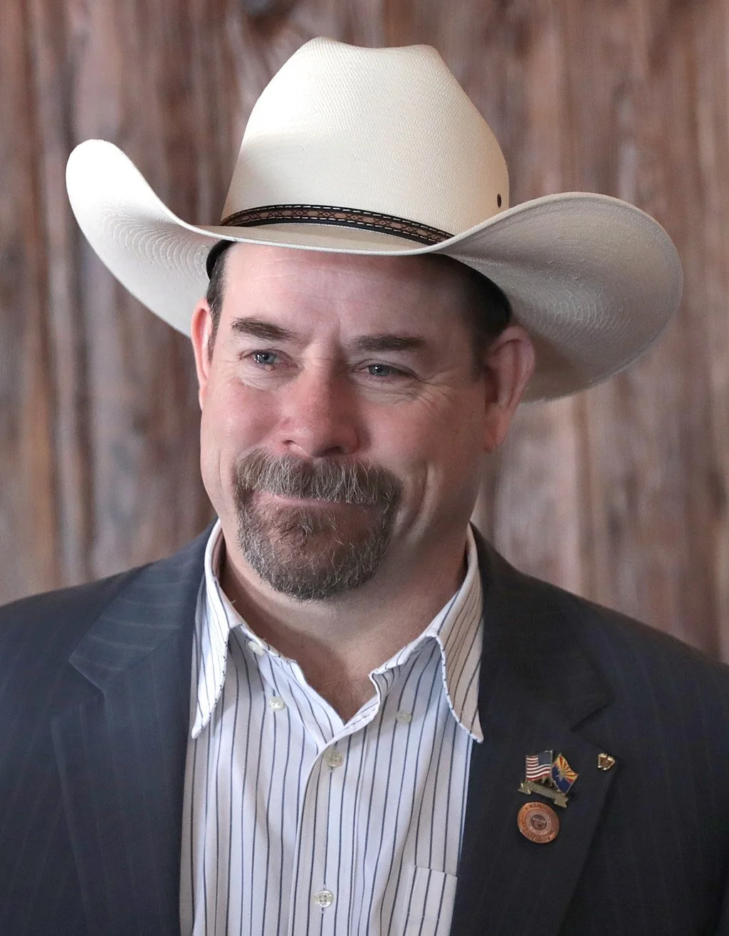 A man wearing a white cowboy hat, a dark pinstripe suit, and a striped dress shirt. He has a beard and mustache and is smiling slightly. He has pins and badges on his suit lapel, including an American flag and other symbols. The background is a wooden wall.