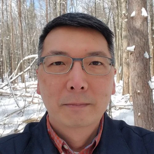 A man wearing glasses and a dark jacket taking a selfie in a snowy forest.