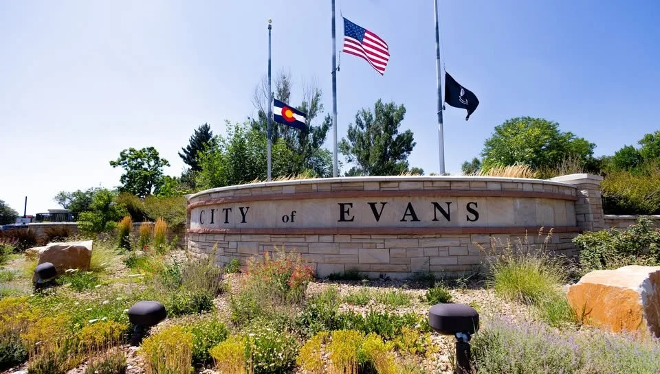 Stone sign reading 'City of Evans' with three flags flying above: the United States flag, the Colorado state flag, and a black flag, with greenery and trees in the background.