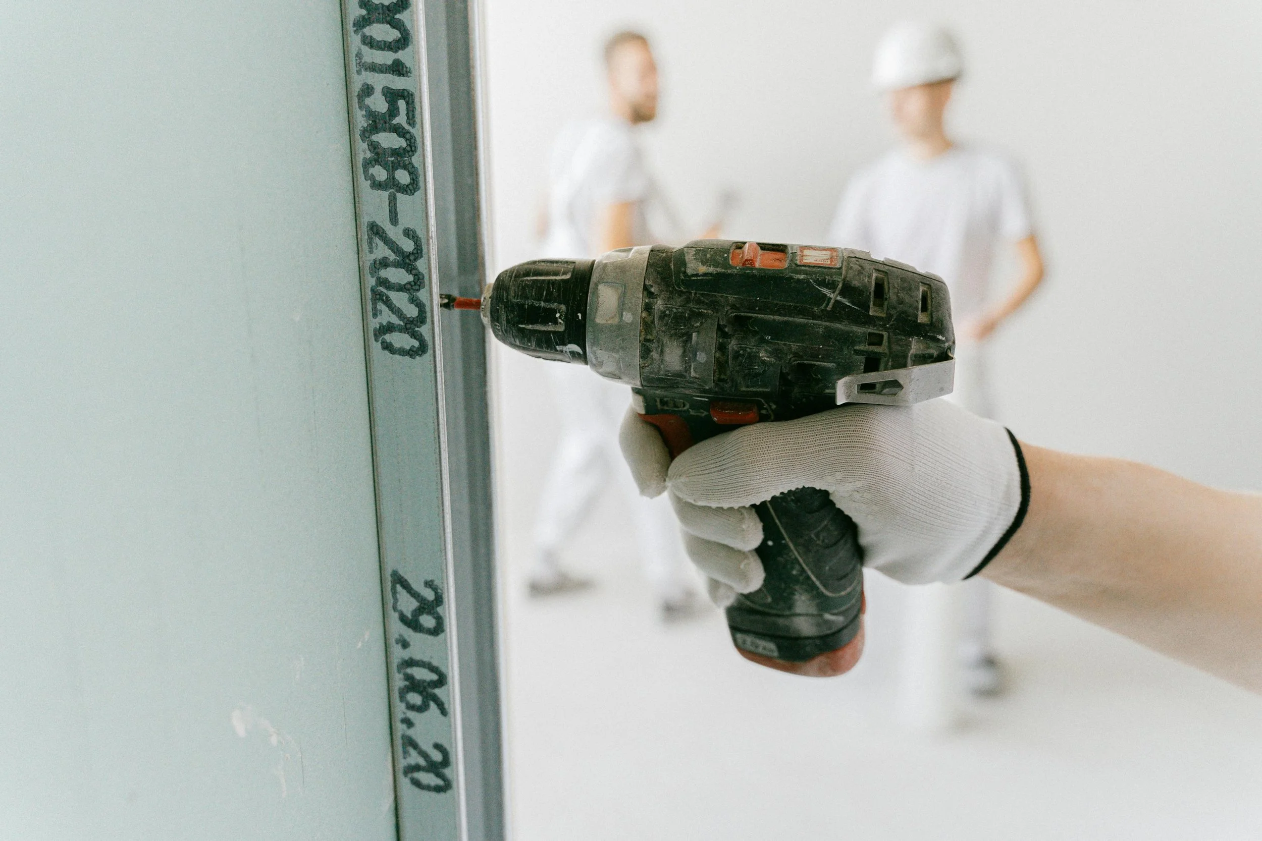 Close-up of a gloved hand using a cordless drill to insert a screw into a drywall. Two people in construction uniforms and safety helmets are blurred in the background.