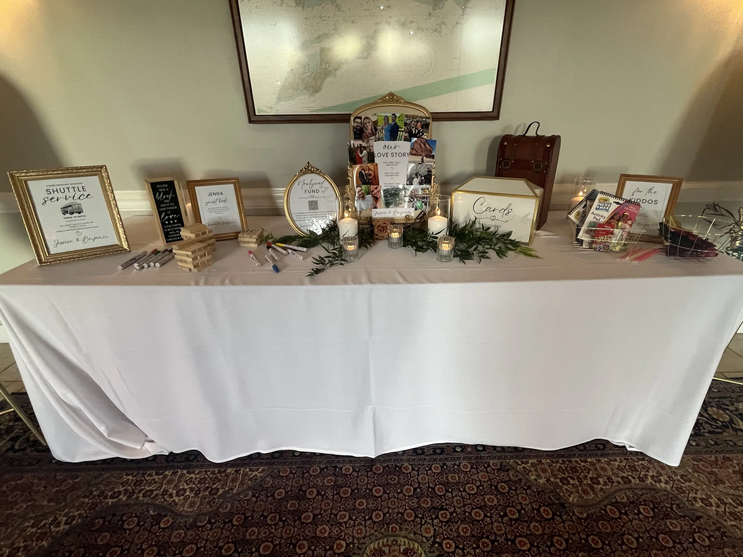 Wedding welcome table with framed signs, pens, coasters, small candles, greenery, and an assortment of cards, messages, and small gift items displayed on a long white table cloth.