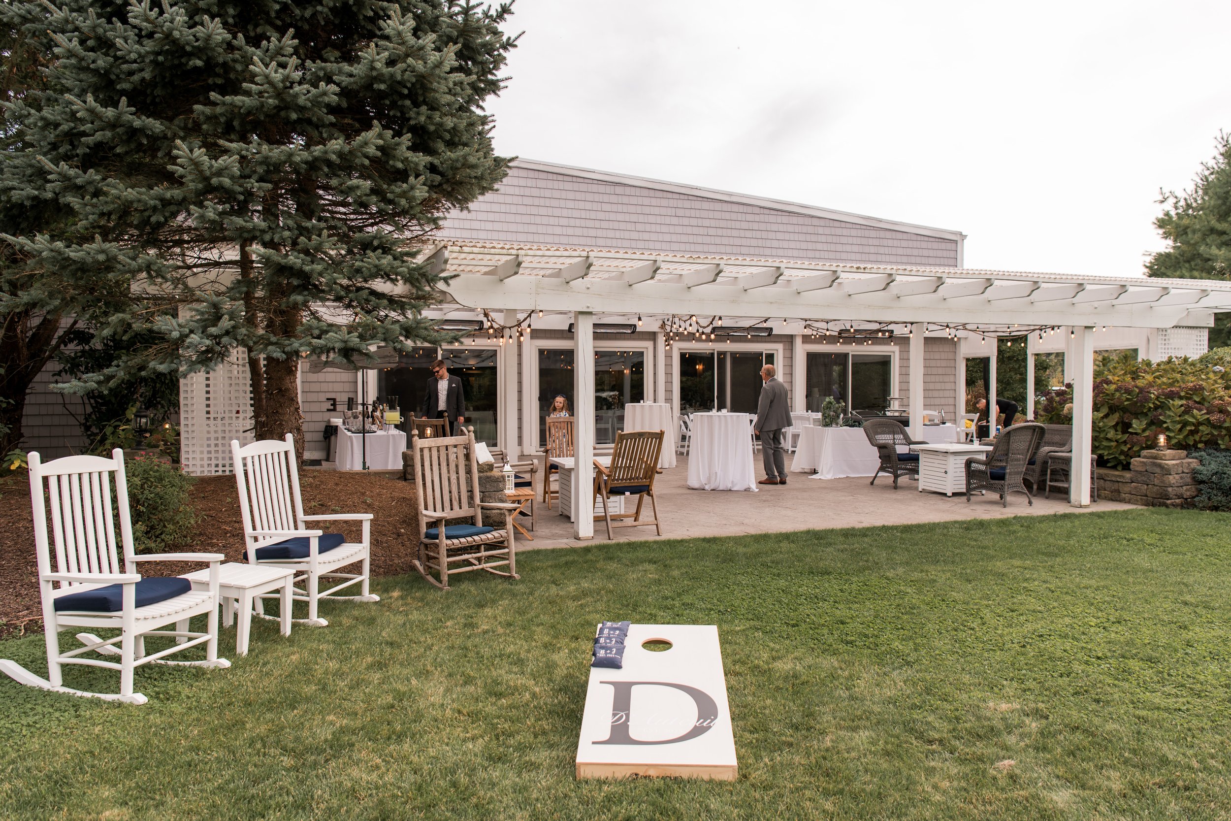 Outdoor party setup under a white pergola, with white and wooden chairs, tables, and string lights, on a green lawn next to a house with gray siding and large windows.